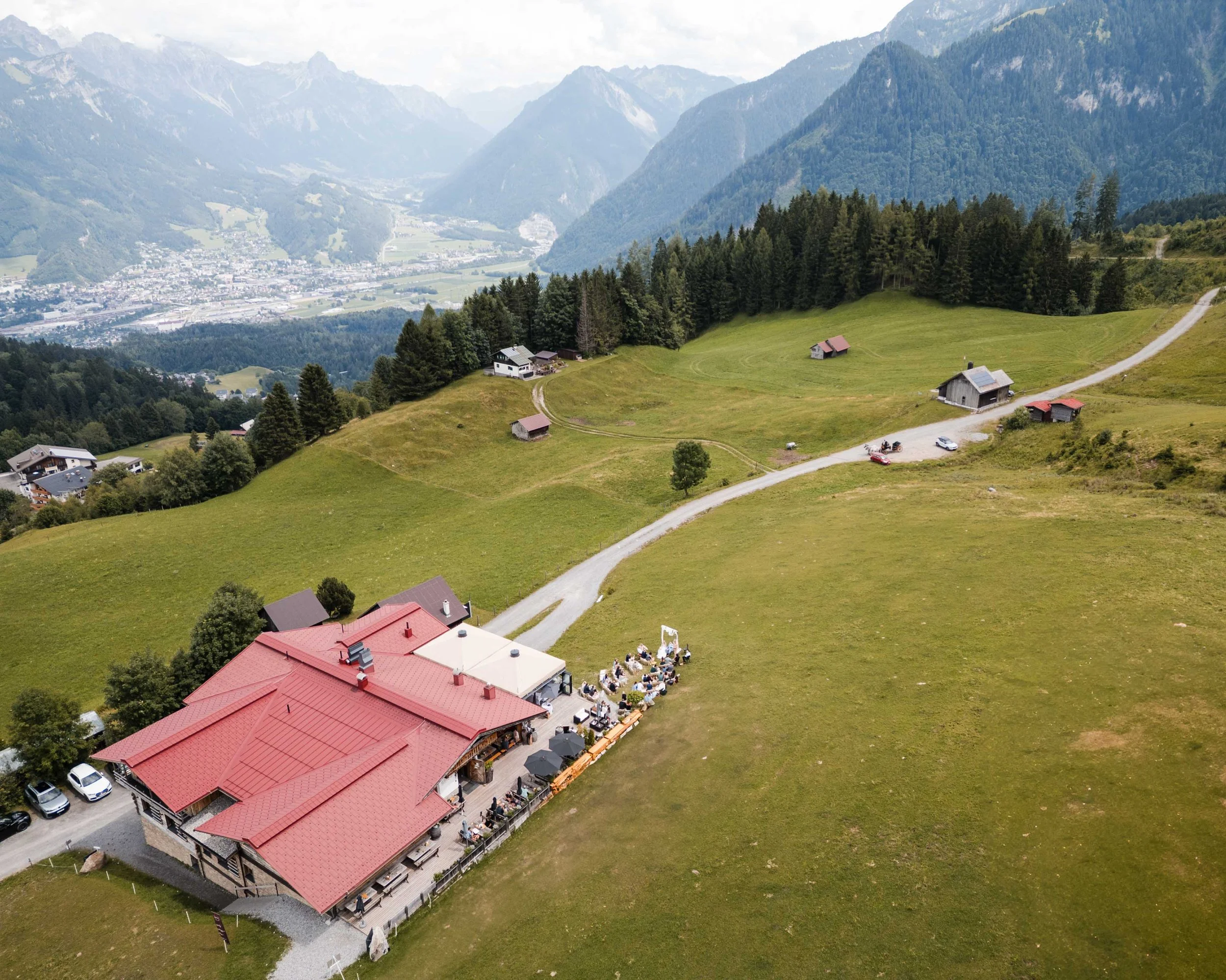 Luftaufnahme eines Restaurants mit rotem Dach in einer Bergregion, umgeben von grünen Wiesen und einzelnen Häusern, mit Bergen im Hintergrund.
