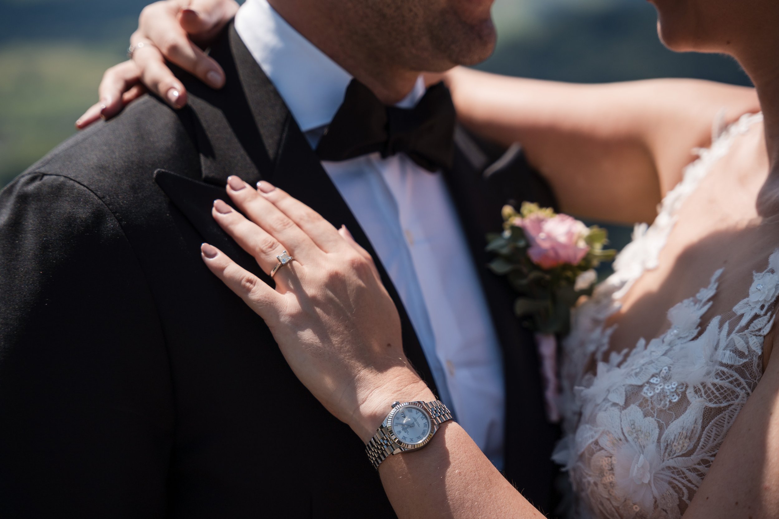 Nahaufnahme eines Brautpaares bei der Eheschließung, Fokus auf die Hände mit Eheringen, der Braut trägt eine Uhr, der Hintergrund ist unscharf.