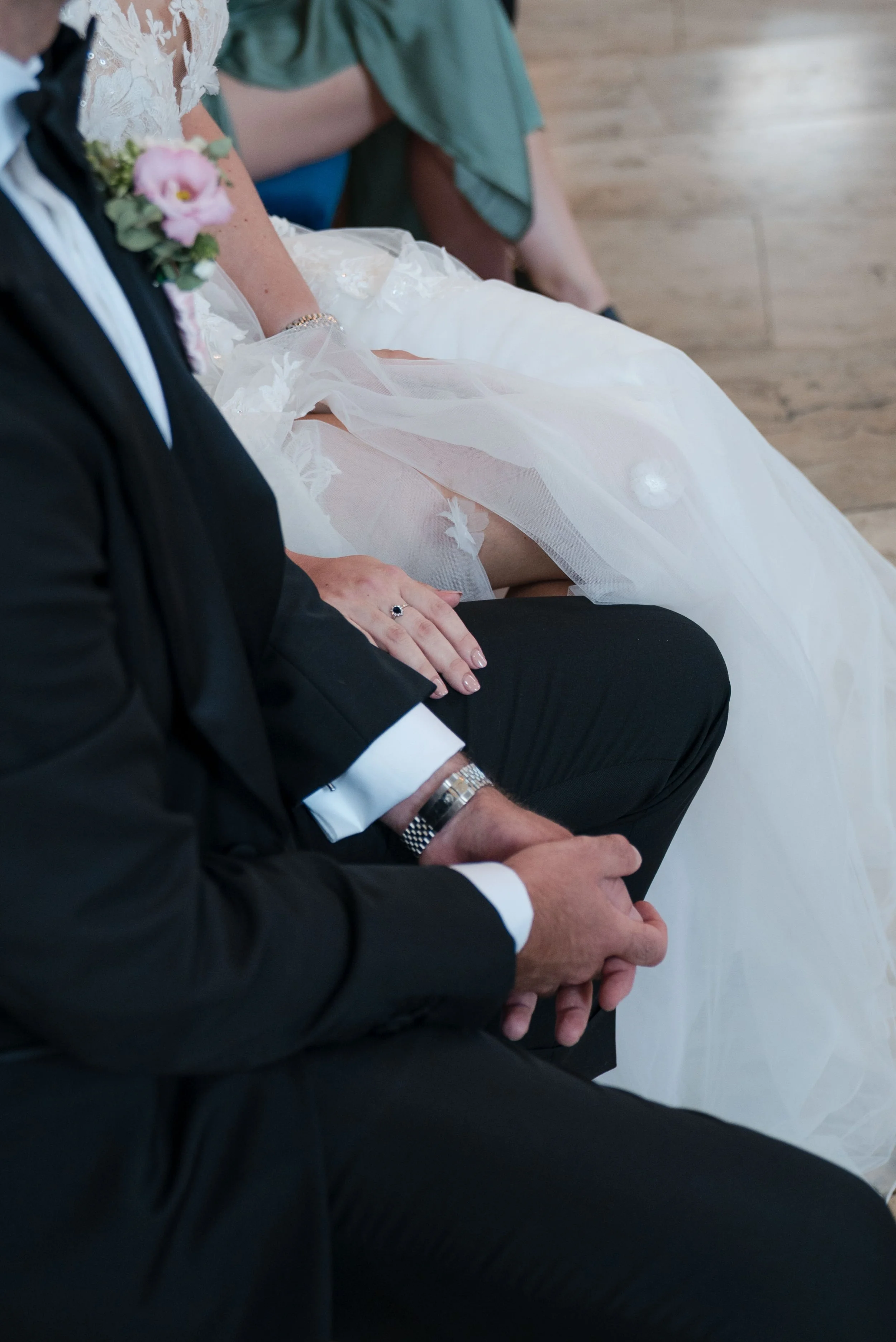 Nahaufnahme von einer Hochzeitszeremonie mit einem Brautpaar, bei dem der Fokus auf den Händen und Kleidung liegt, einschließlich eines weißen Brautkleids und eines schwarzen Anzugs.