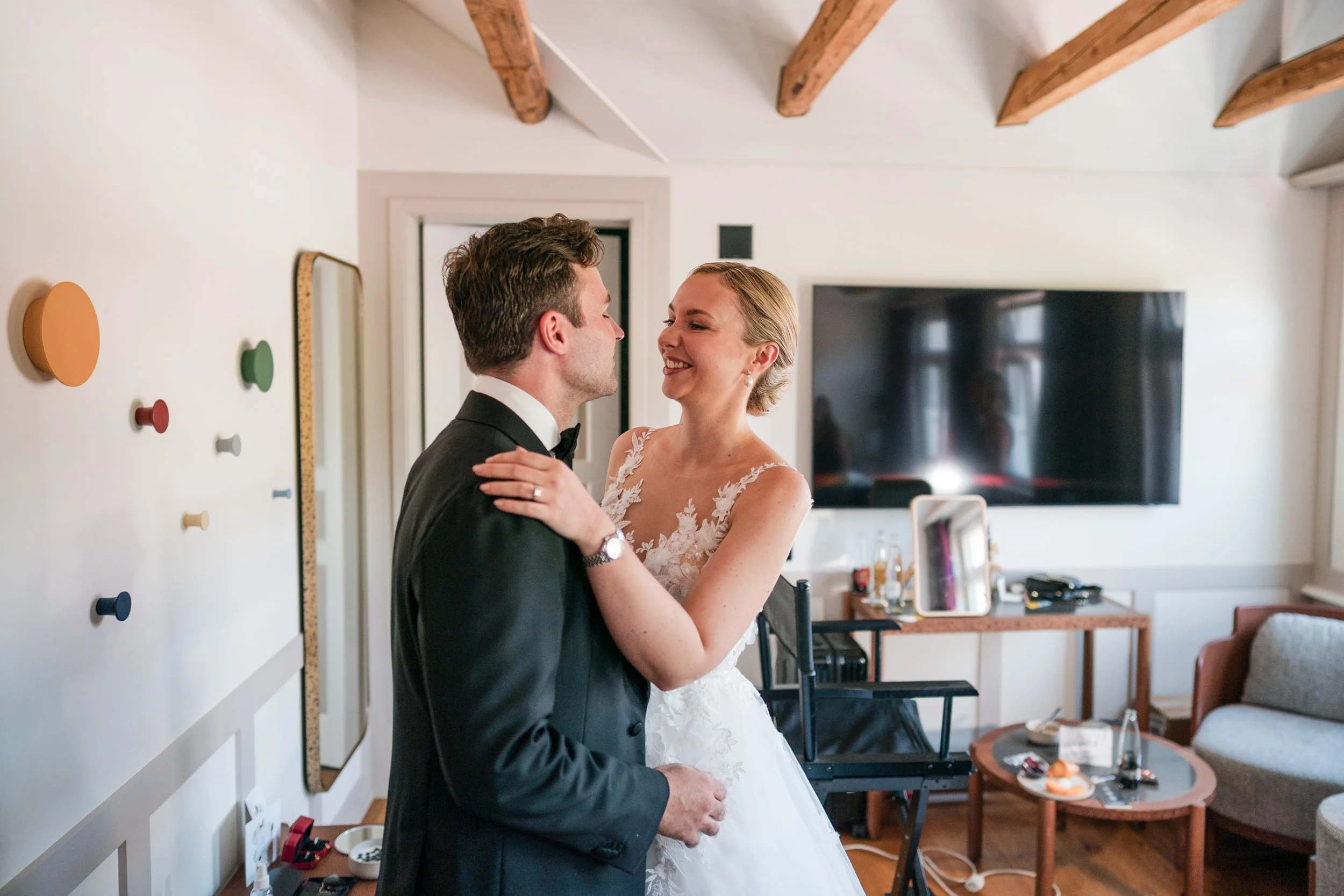 Ein Brautpaar in einem eleganten Raum, das sich verträumt ansieht. Der Mann trägt einen schwarzen Anzug und eine Fliege, die Frau ein weißes Brautkleid mit Spitze.