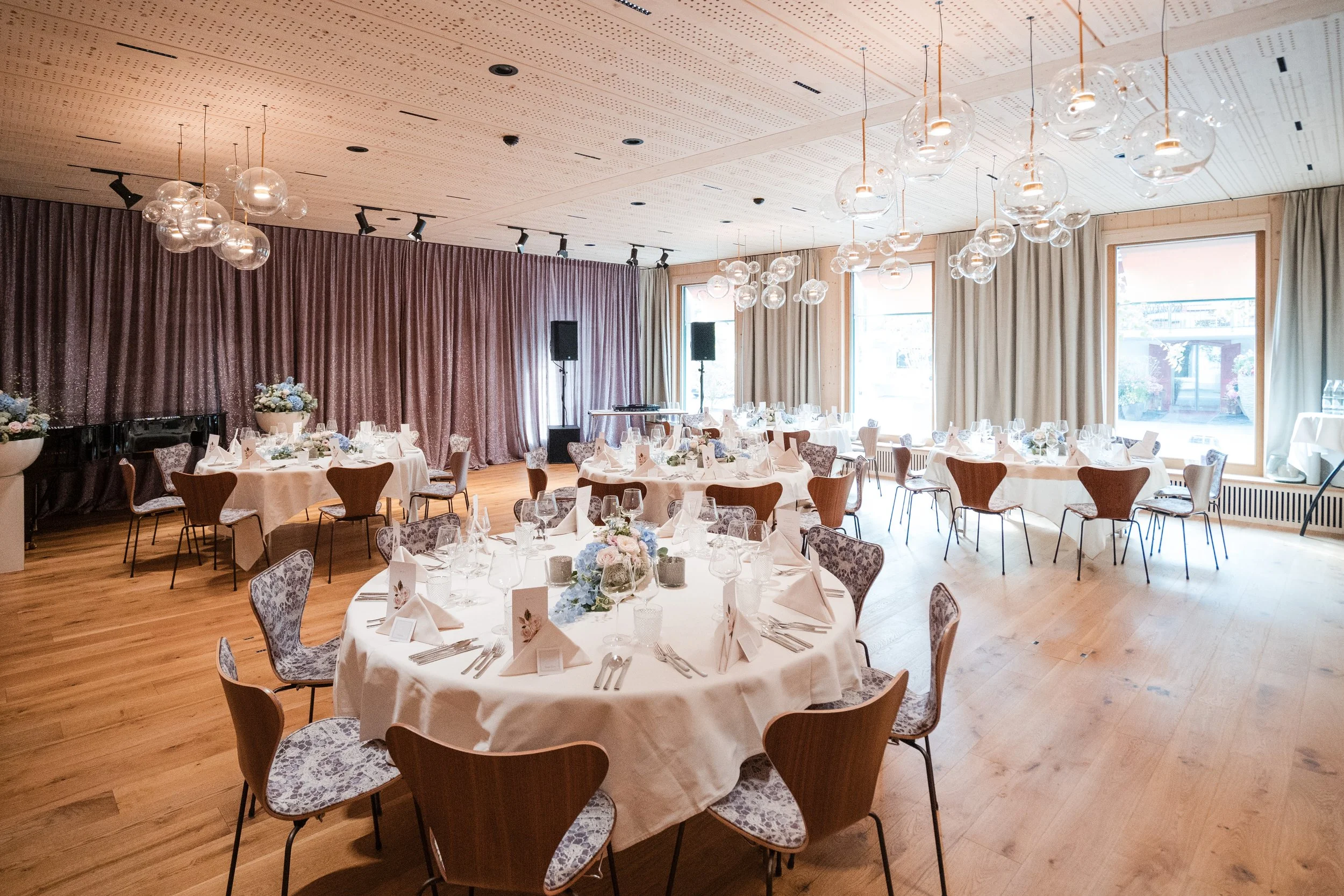 Elegant gedeckter Veranstaltungsraum mit runden Tischen, Blumenarrangements, Kerzen, Weingläsern und Besteck, große Fenster mit Vorhängen, moderne Beleuchtung, Holzfußboden.