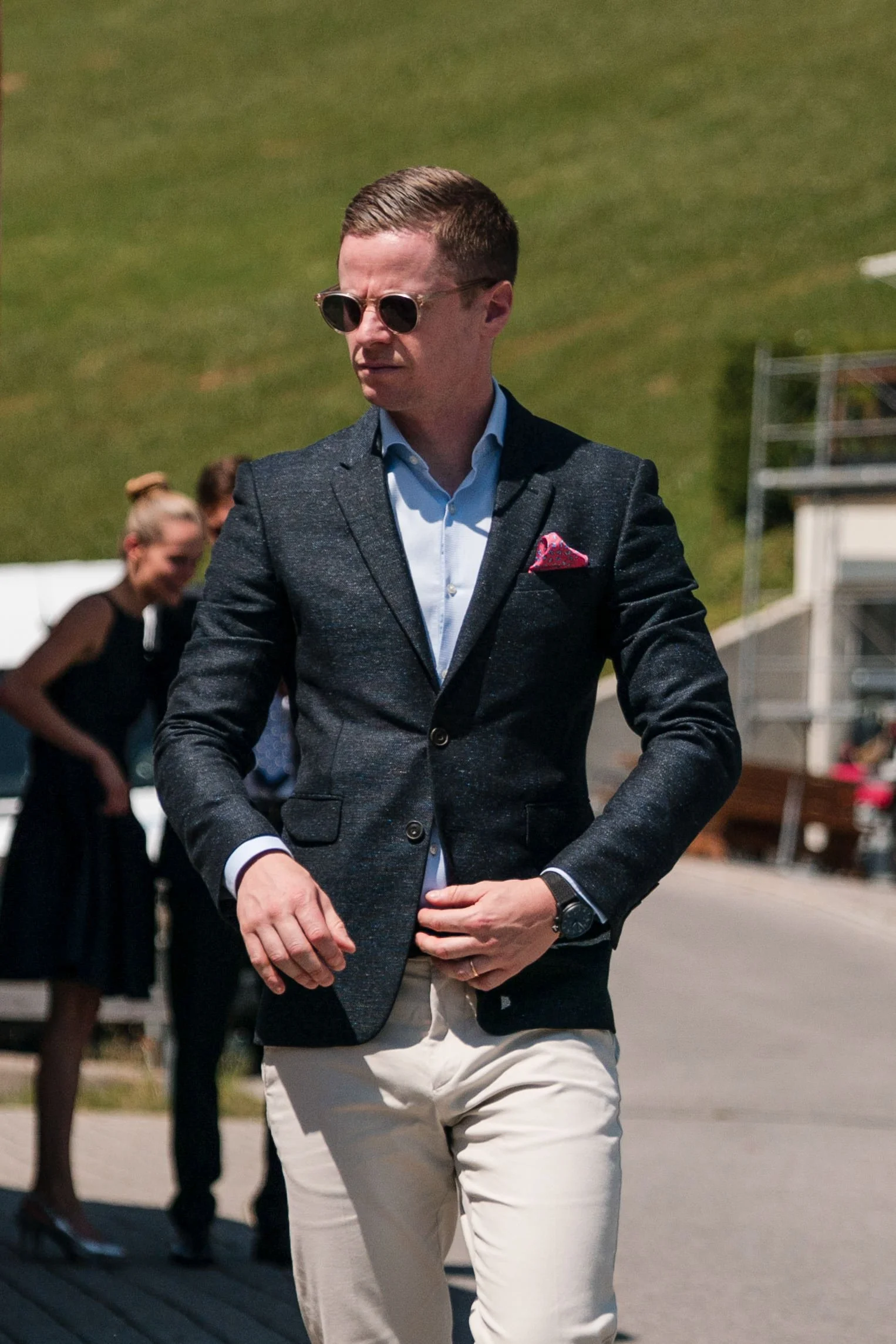 Ein Mann in dunkler Jacke, hellblauem Hemd, heller Hose und Sonnenbrille steht draußen bei sonnigem Wetter.