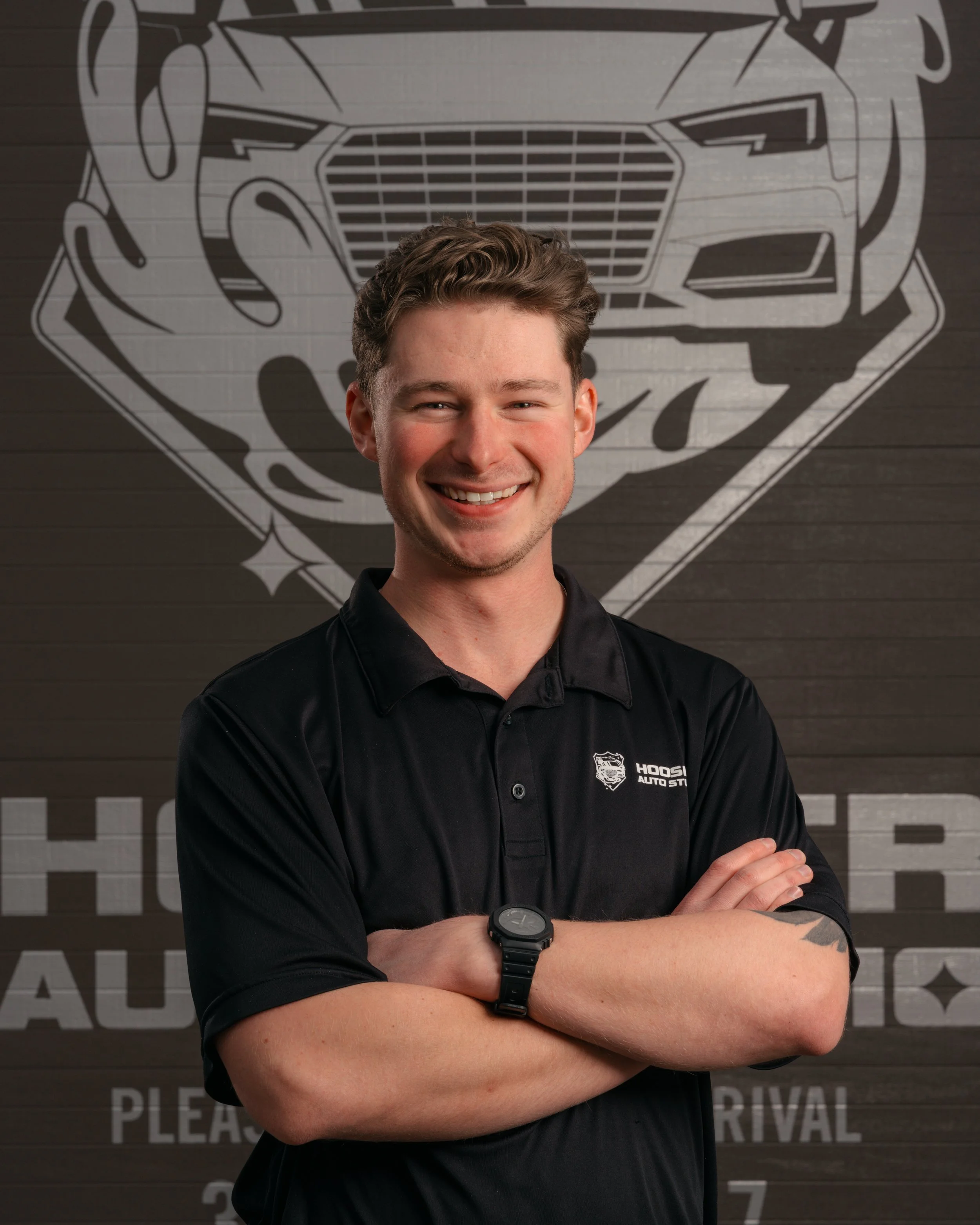 A young man with light brown hair smiling and standing with arms crossed in front of a wall with a large logo.  Text on the shirt says 'Hoosier Auto Studio'. Modern professional headshot for personal branding and online presence in Indianapolis
