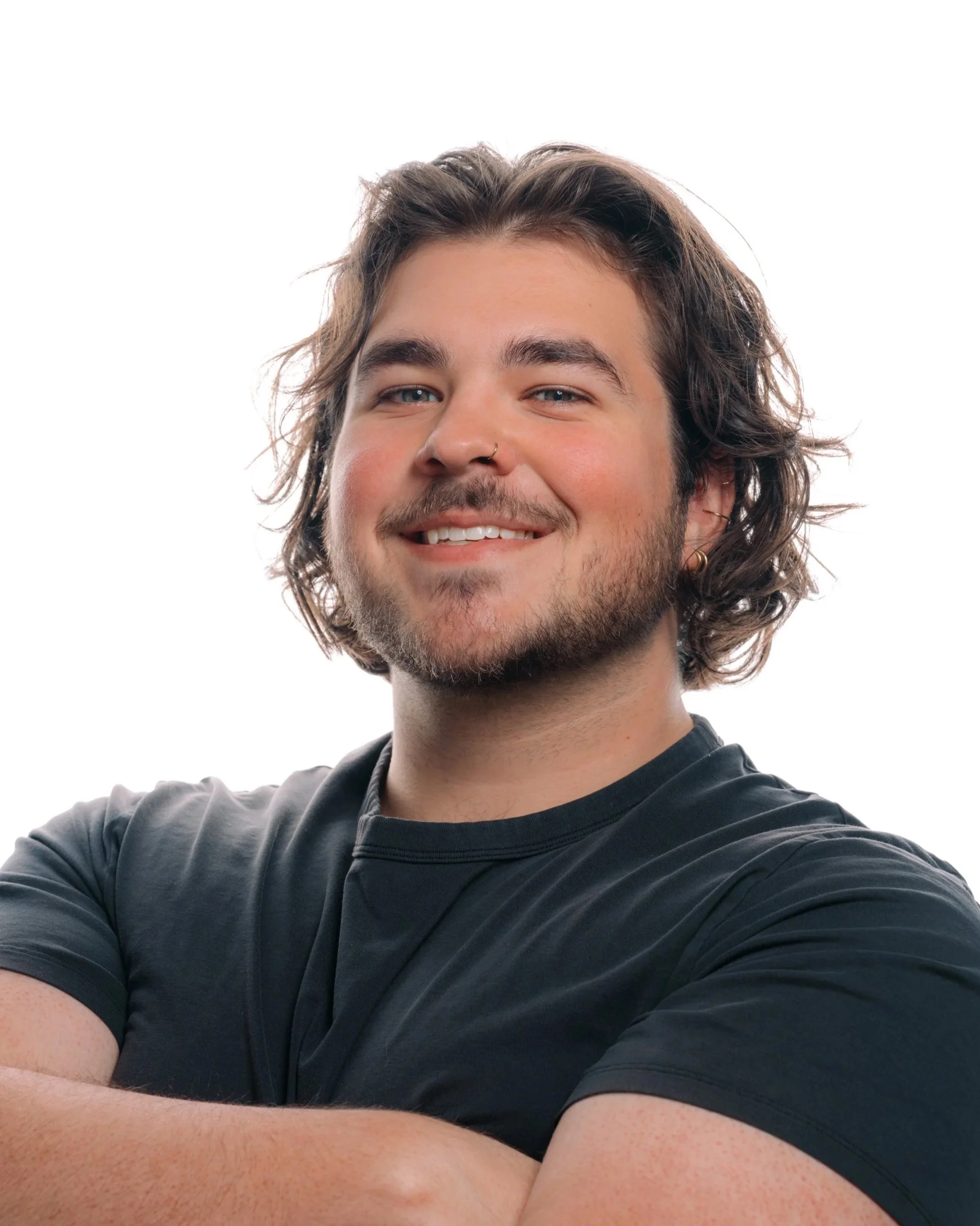 Portrait of a smiling young man with wavy brown hair, blue eyes, and facial hair, wearing a black T-shirt and earrings, against a white background. Clean modern headshot for LinkedIn profile Indianapolis photographer