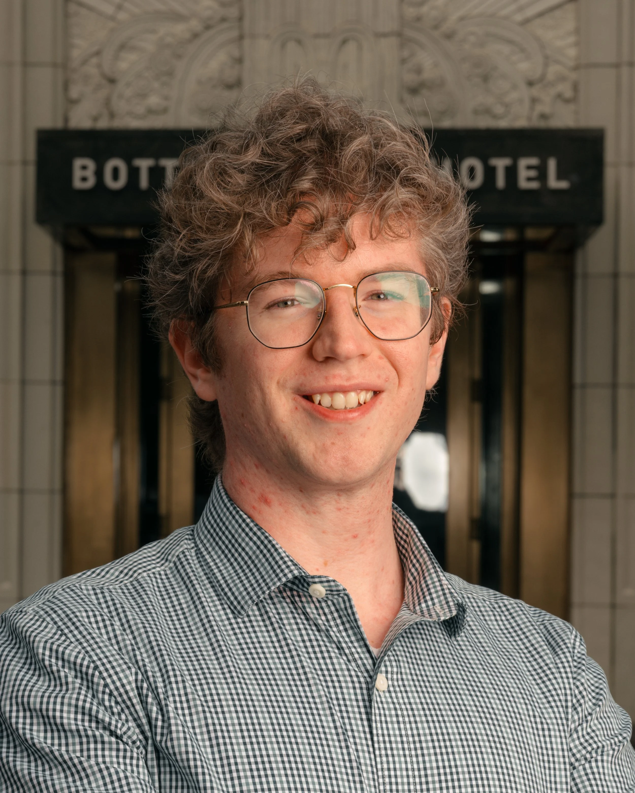 A young man with curly hair, glasses, and a checkered shirt smiling in front of a hotel entrance.