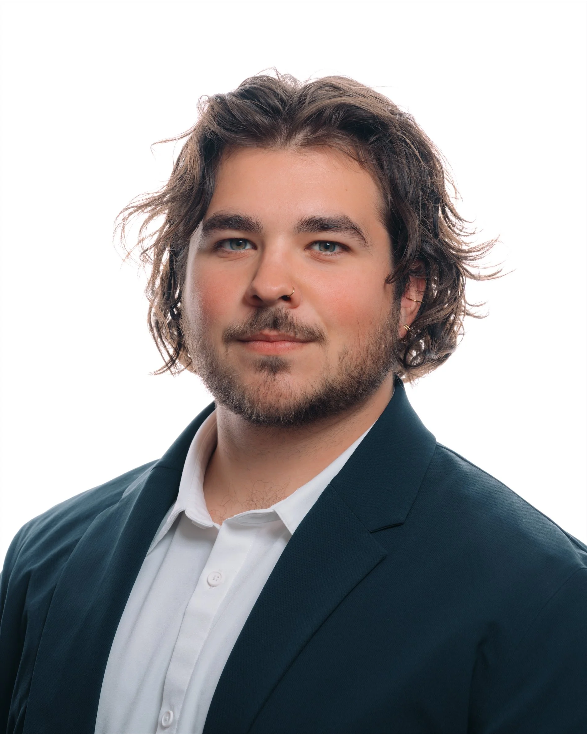 A young man with wavy brown hair, blue eyes, and a beard, wearing a black suit jacket and a white dress shirt, against a white background. Studio-lit headshot for business professional focused on confidence and credibility Indianapolis photography