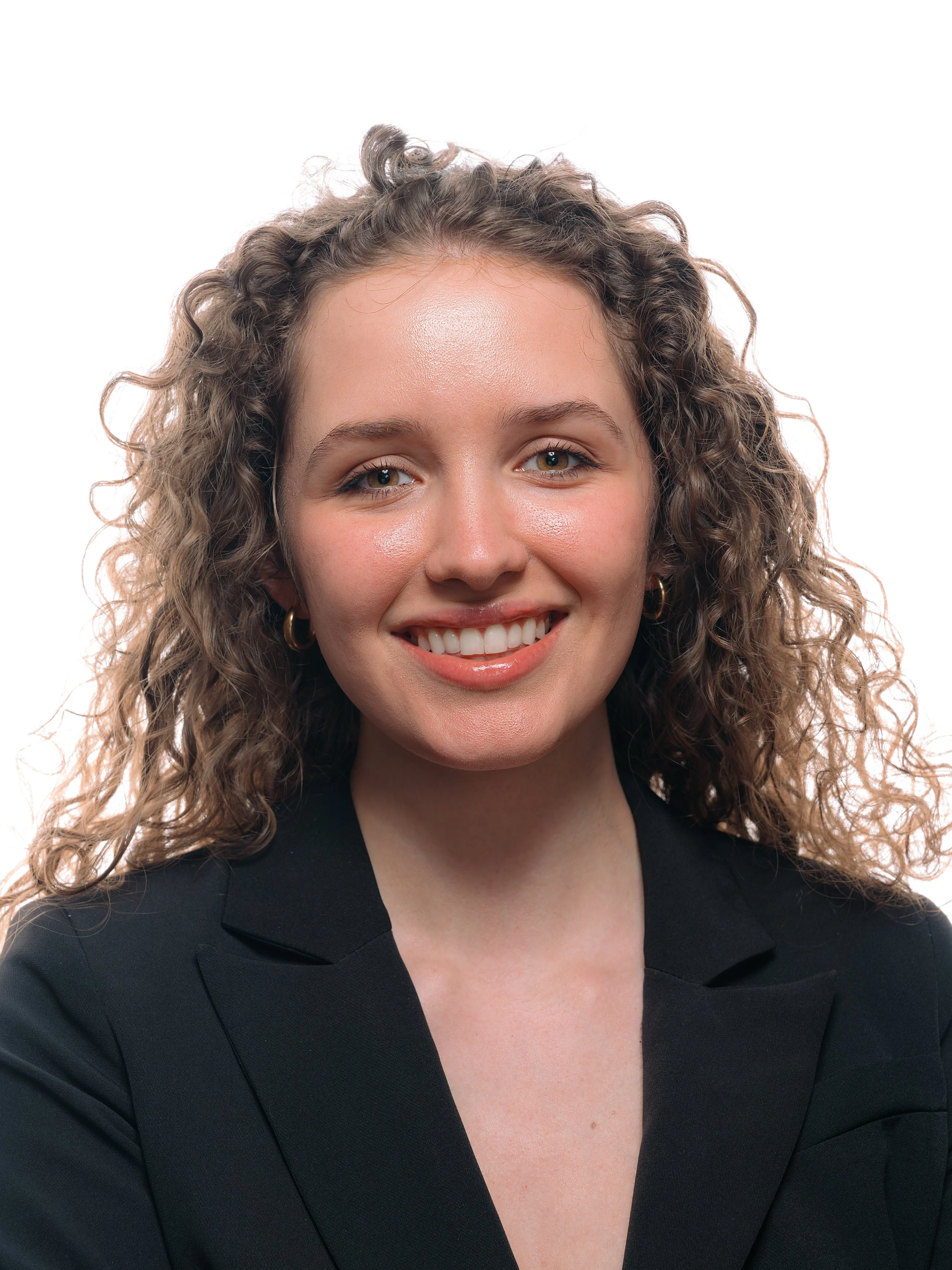 Close-up of a young woman with curly blonde hair, wearing a black blazer and gold hoop earrings, smiling against a white background. Corporate headshot for entrepreneur in Indianapolis photography session