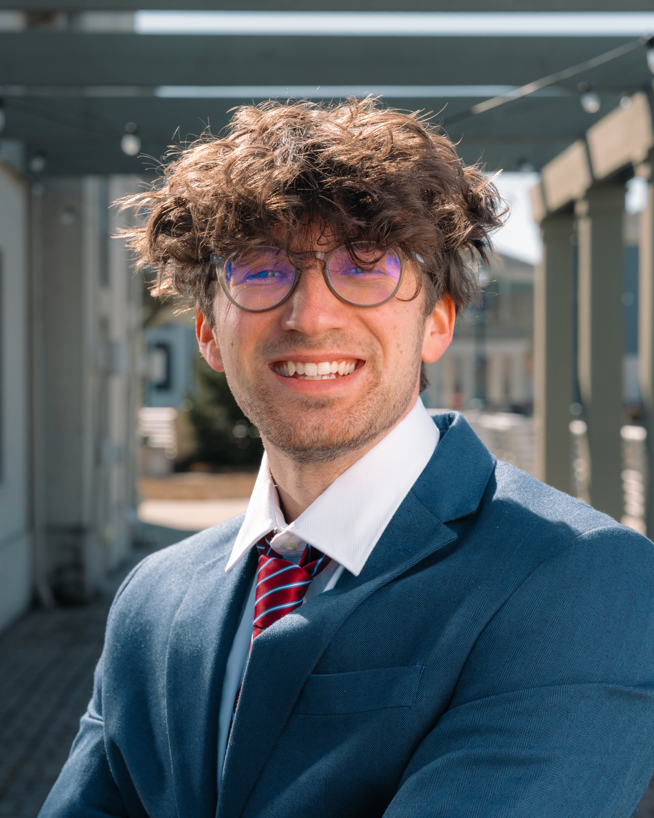 Man in a business suit with curly hair and glasses, smiling outdoors on a sunny day.
