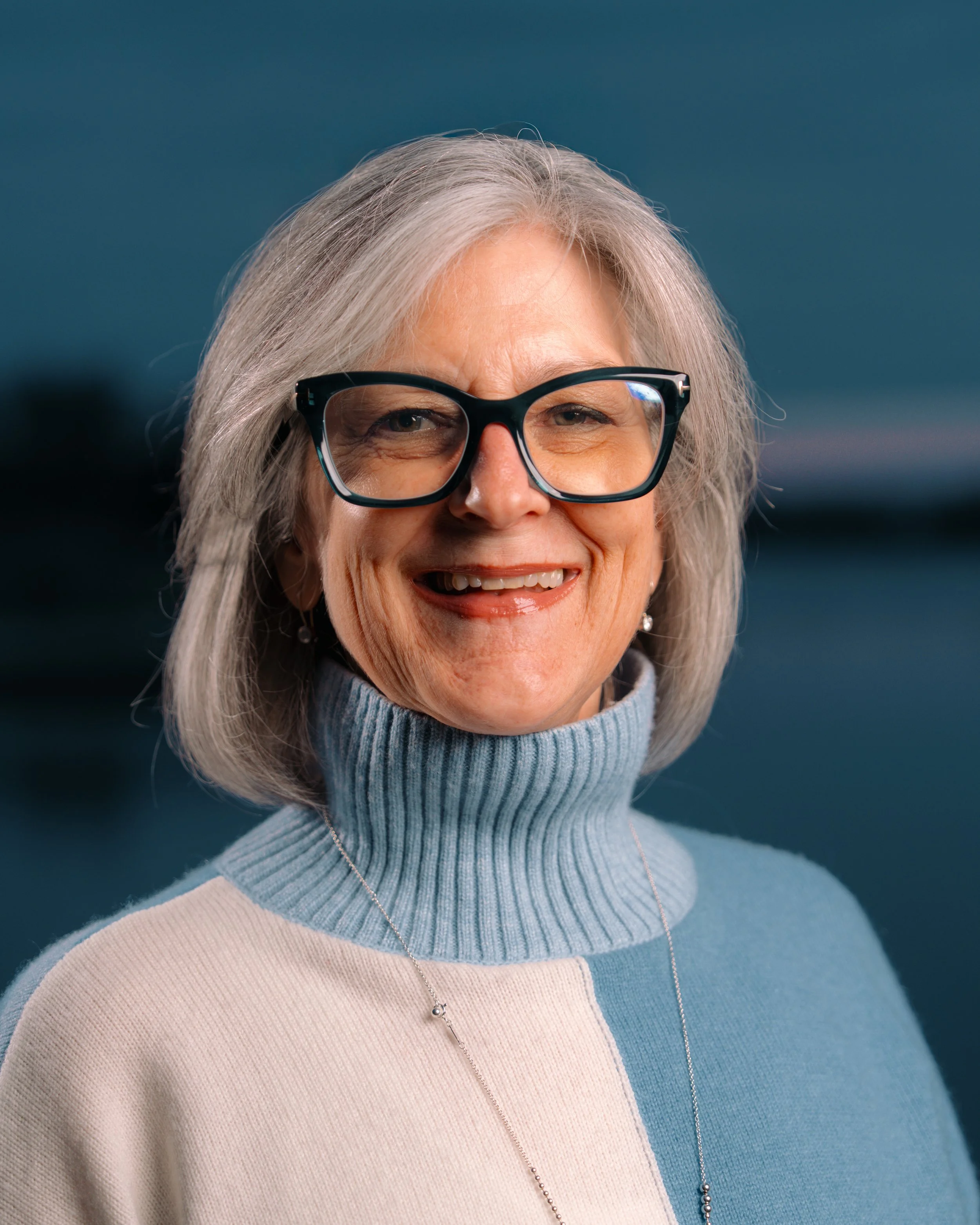 Close-up portrait of a woman wearing black glasses, a turtleneck sweater in light blue and beige, and a delicate necklace, smiling outdoors against a blurred background. Editorial style portrait photography with dramatic lighting Indianapolis