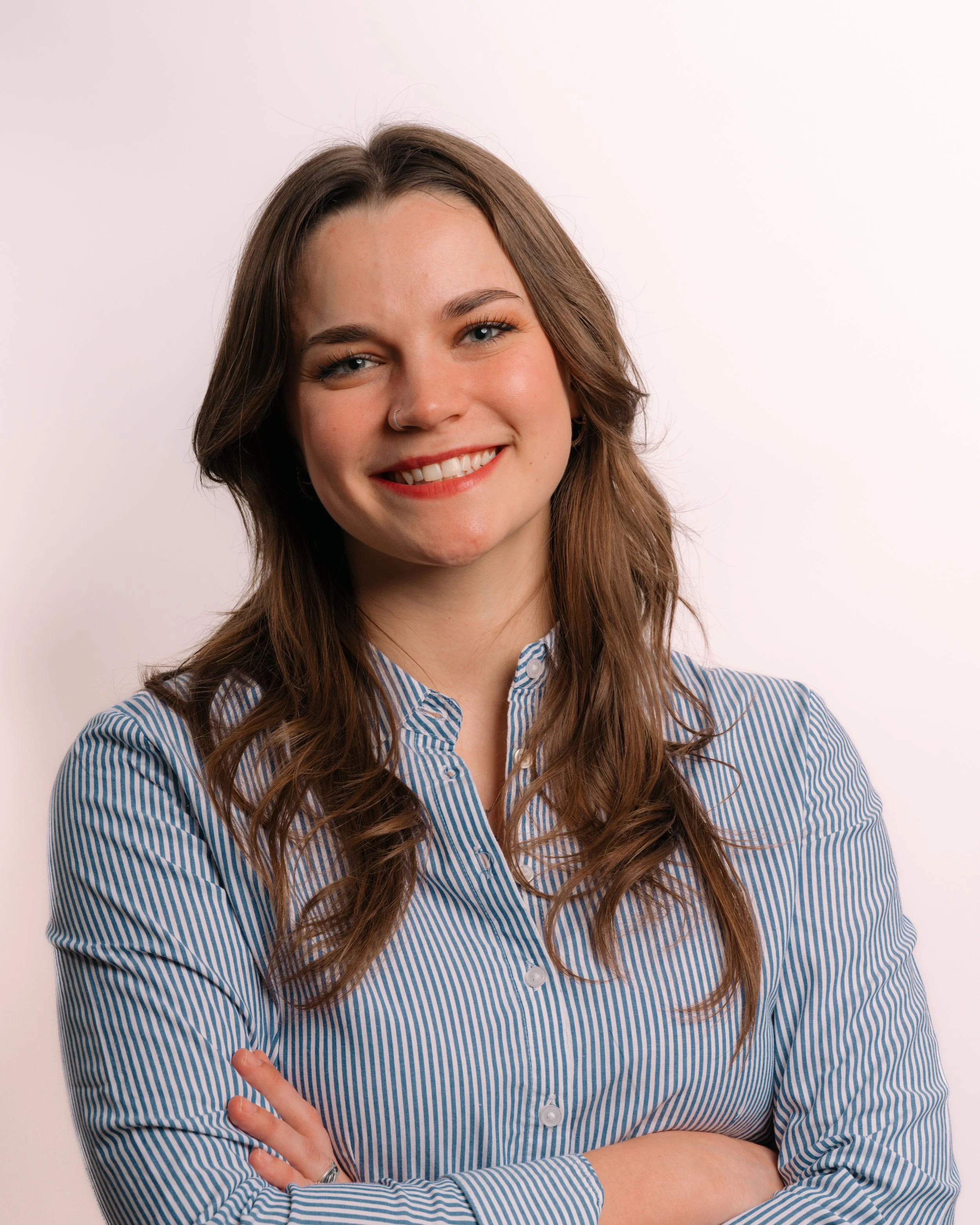 A young woman with long brown hair, blue eyes, and a nose ring, wearing a blue and white striped shirt, smiling with arms crossed, standing against a white background.