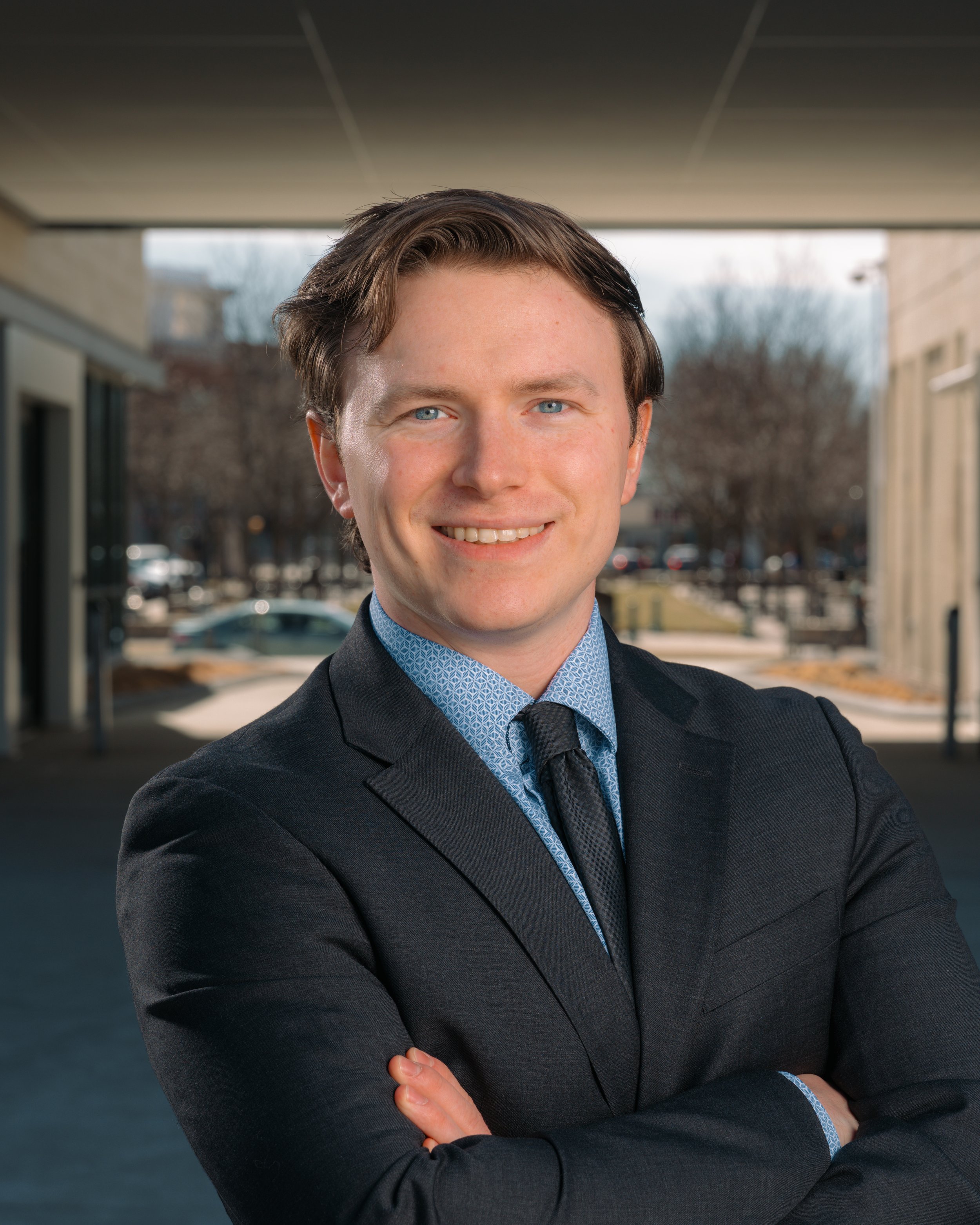 A young man in a dark suit with crossed arms standing outdoors with a modern building and parked cars in the background. Business branding photography session for professional website content