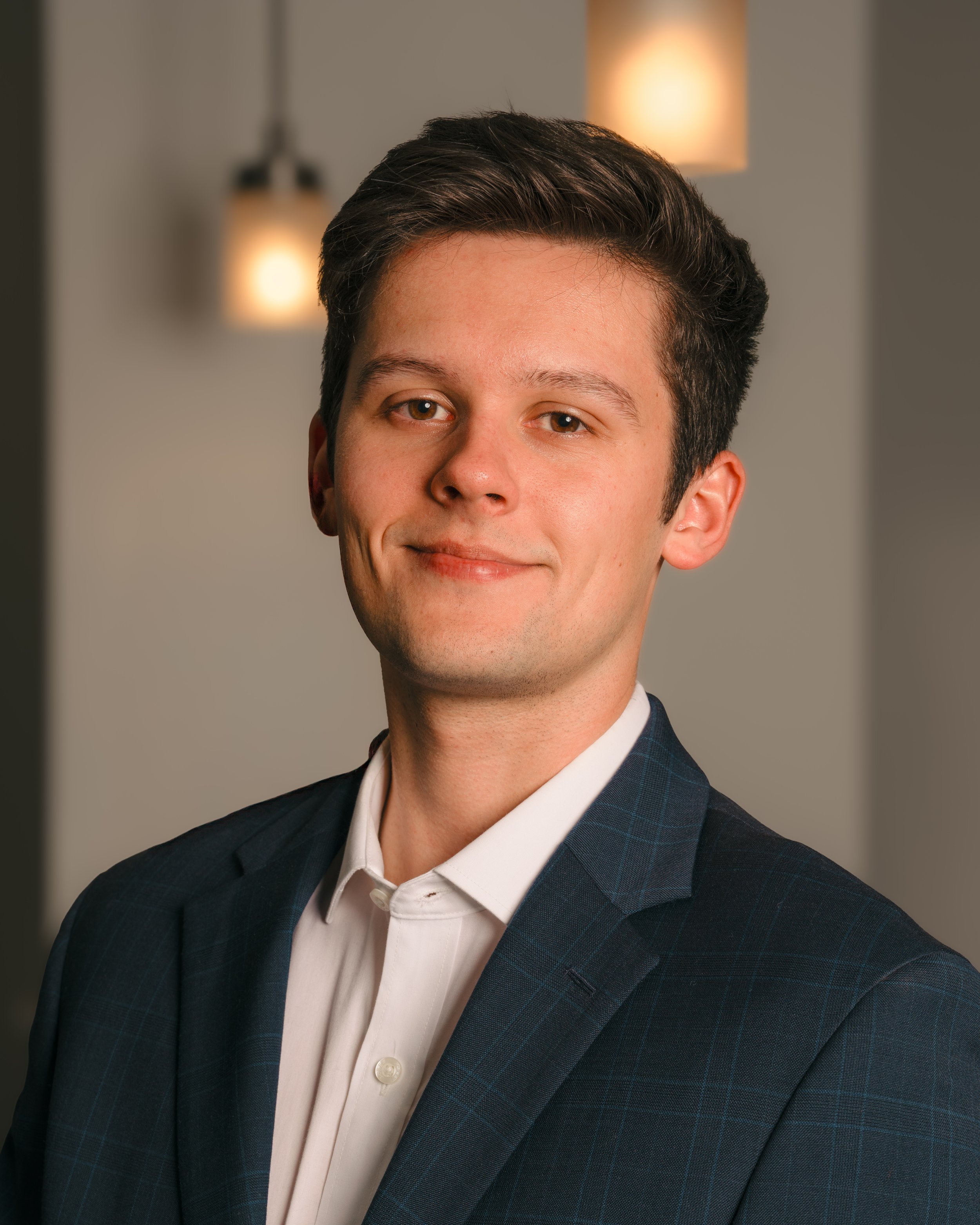 A young man with dark hair, wearing a navy blazer and a white shirt, smiling slightly. Background features soft lighting with pendant lamps.