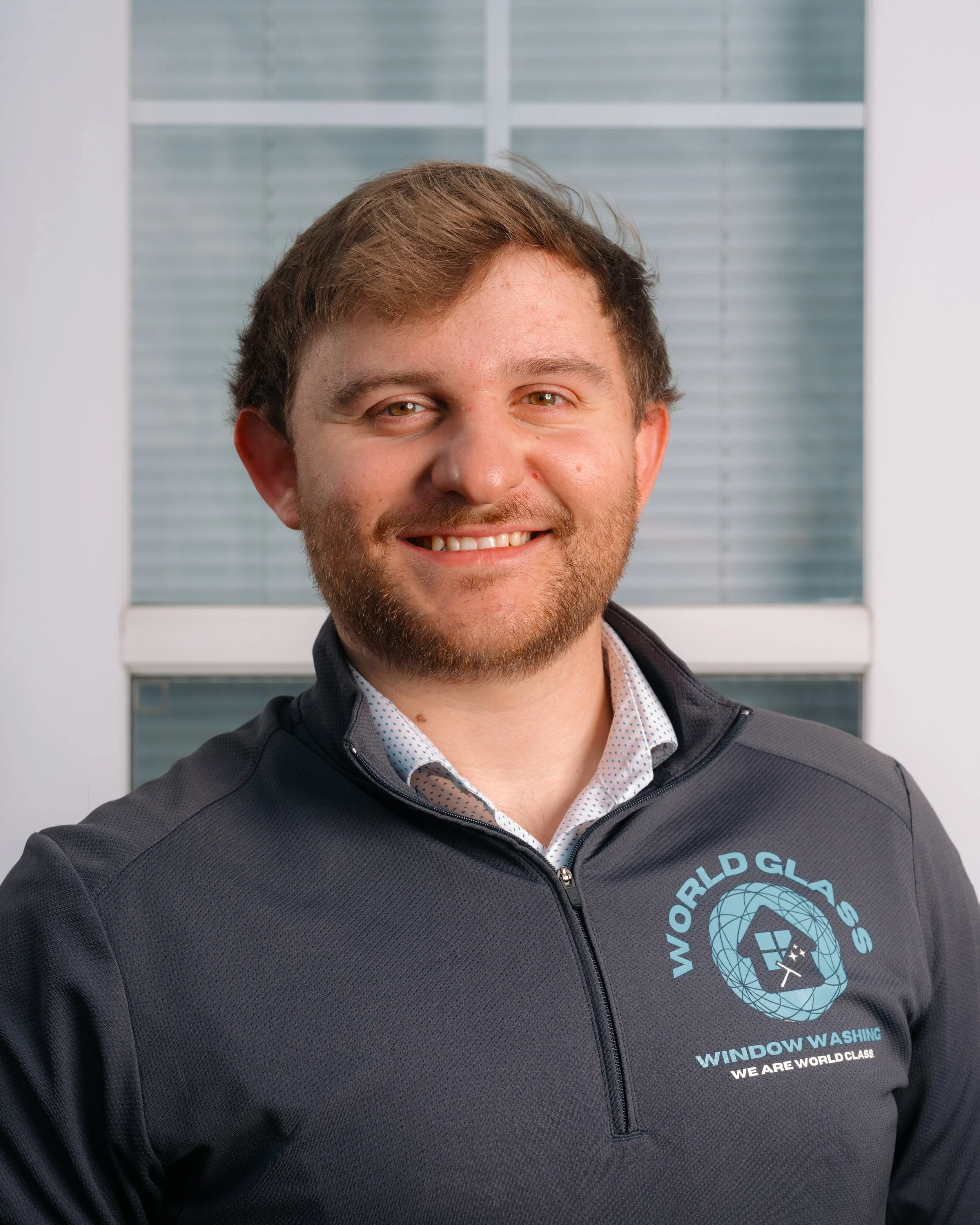 A smiling man with light brown hair and beard wearing a dark zip-up jacket with a logo that says 'World Glass' and 'Window Washing' in front of a window.