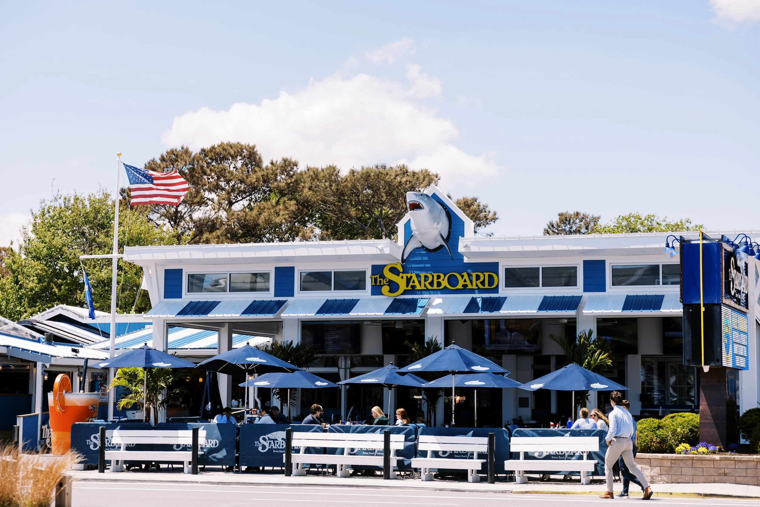 Exterior of The Starboard restaurant with blue and white decorations, American flag, outdoor seating, umbrellas, and people dining.
