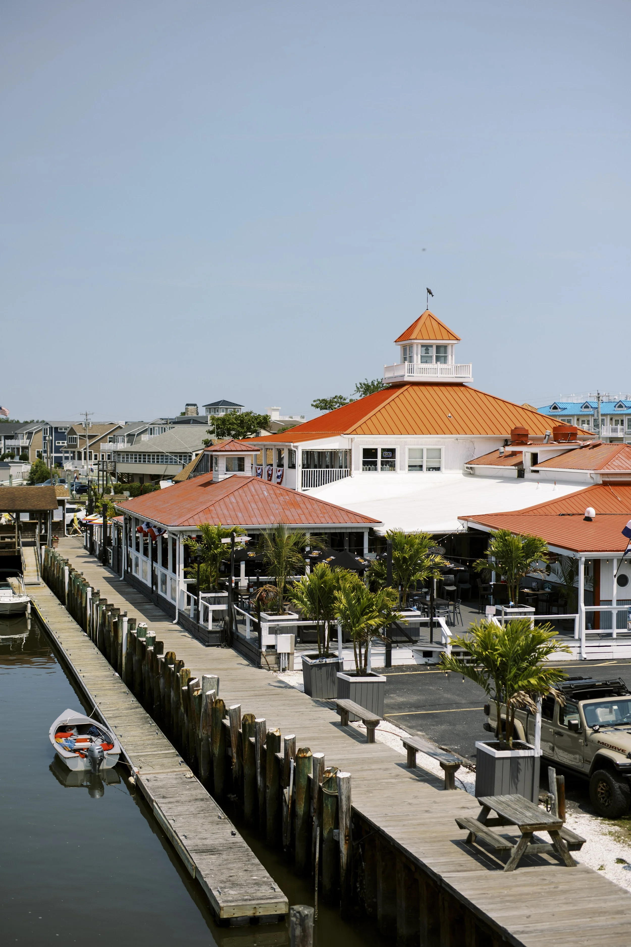 View of a waterfront restaurant with a pier, a boat docked, a large building with a tower and orange roof, palm trees, and residential buildings in the background under a clear blue sky.