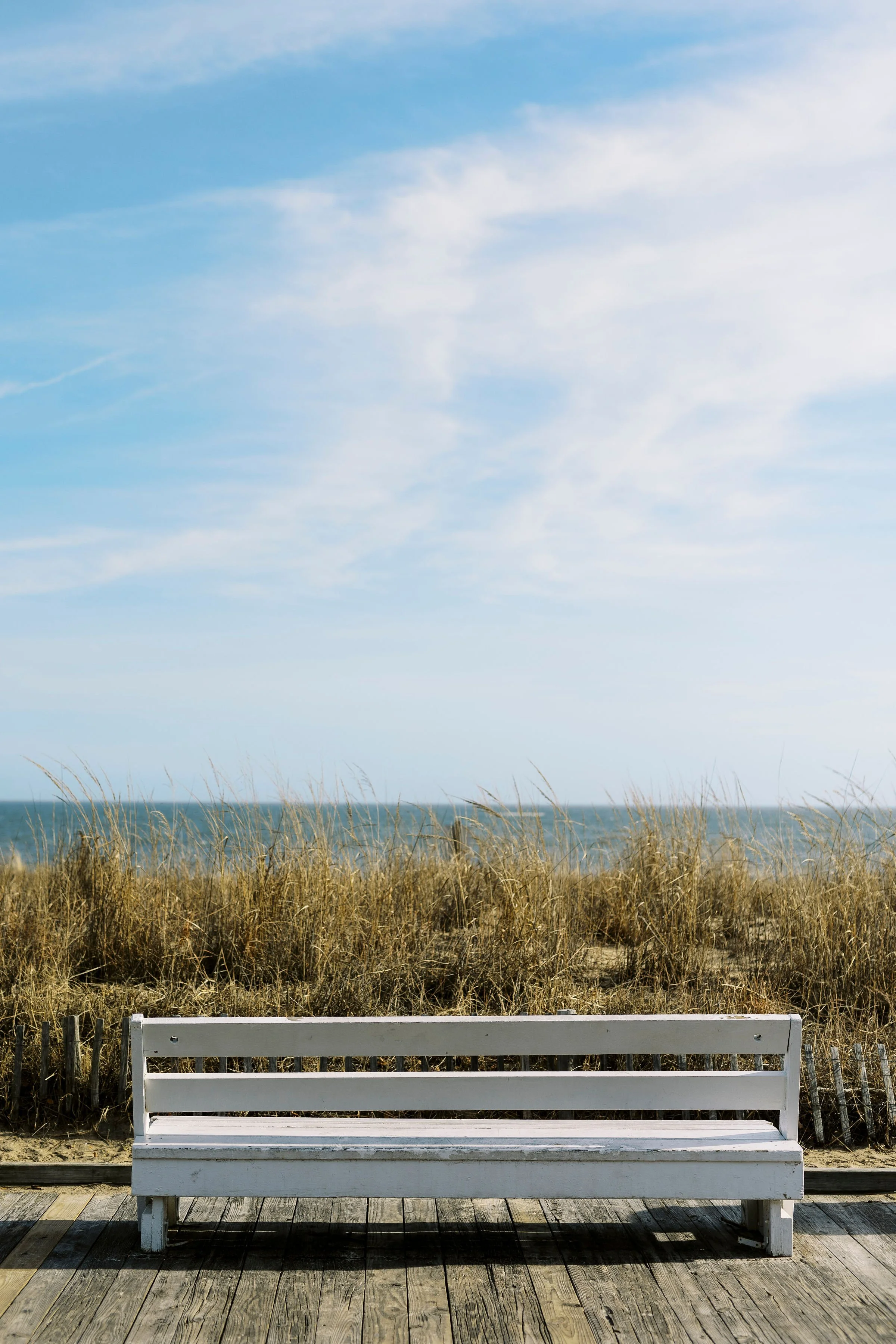 White wooden bench on a wooden deck facing a beach with tall grasses and dunes, overcast sky.