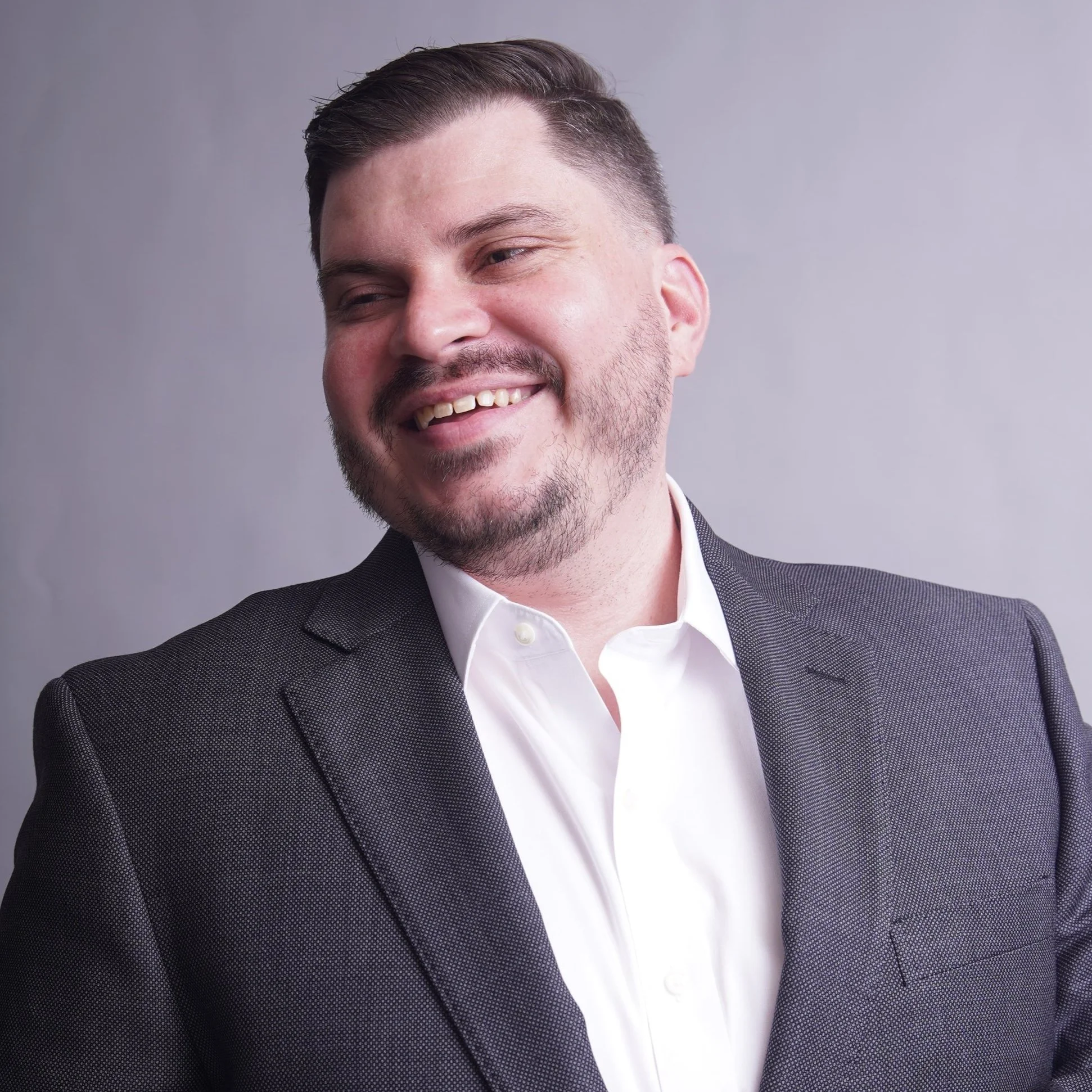 A man with short hair, a beard, and a mustache wearing a dark suit jacket and white dress shirt, smiling and looking to the side.