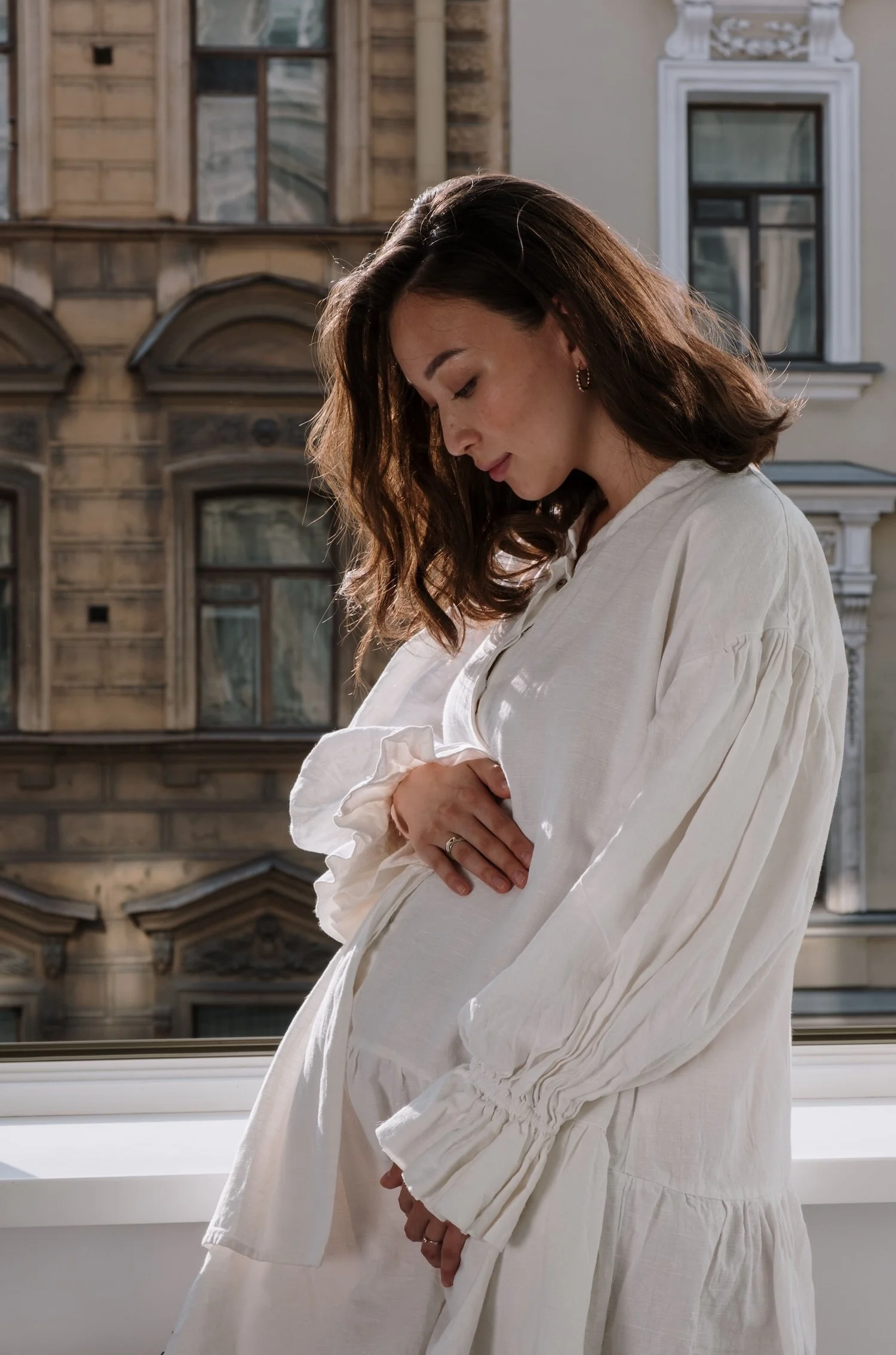 A pregnant woman in a white dress looks down at her belly with a pensive expression. Maternal mental health support Washington DC matters, and pregnancy therapy Washington DC with a psychodynamic therapist in Washington, DC offers space to explore.