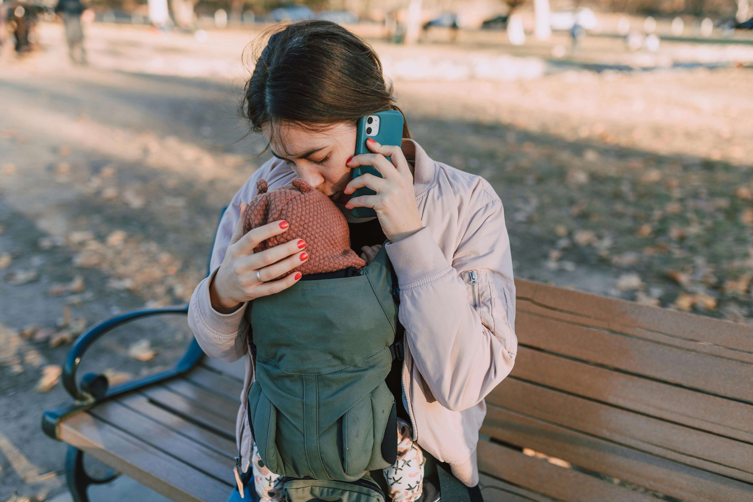 Woman sitting on a park bench talking on her phone while kissing her baby’s head in a carrier, representing complex emotions addressed in postpartum therapy for new moms in Washington, DC.