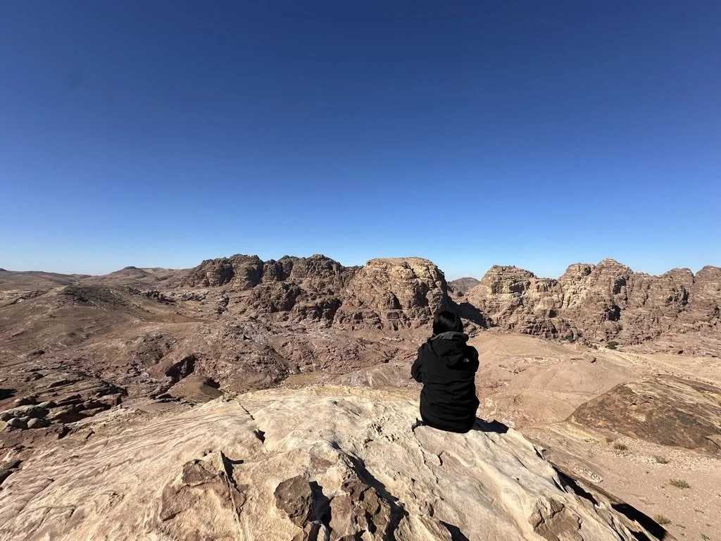 A person wearing a backpack walking through desert sand toward large rock formations under a clear blue sky.