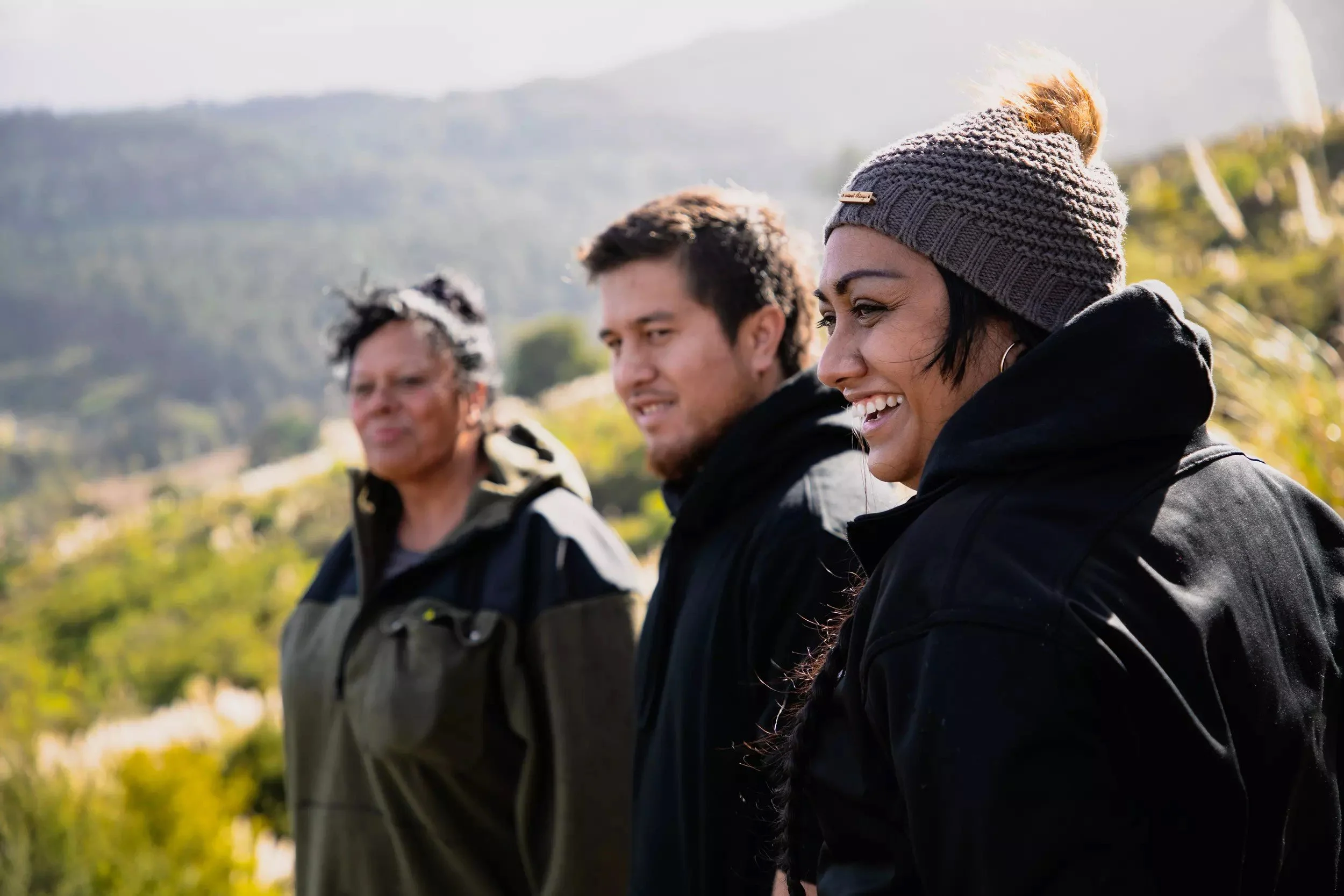 Three people outdoors, smiling and looking to the side, with a scenic background of hills and trees, dressed in casual jackets and a knit hat.