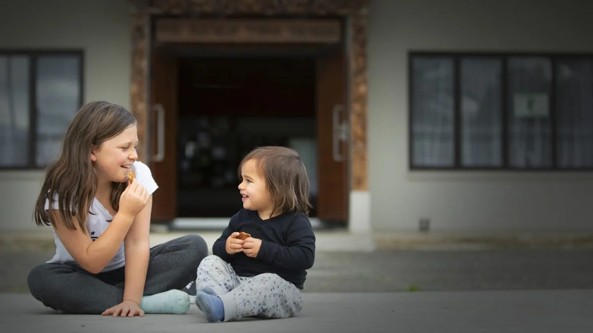 Two young girls sitting on the sidewalk outside a building, laughing and sharing a snack.