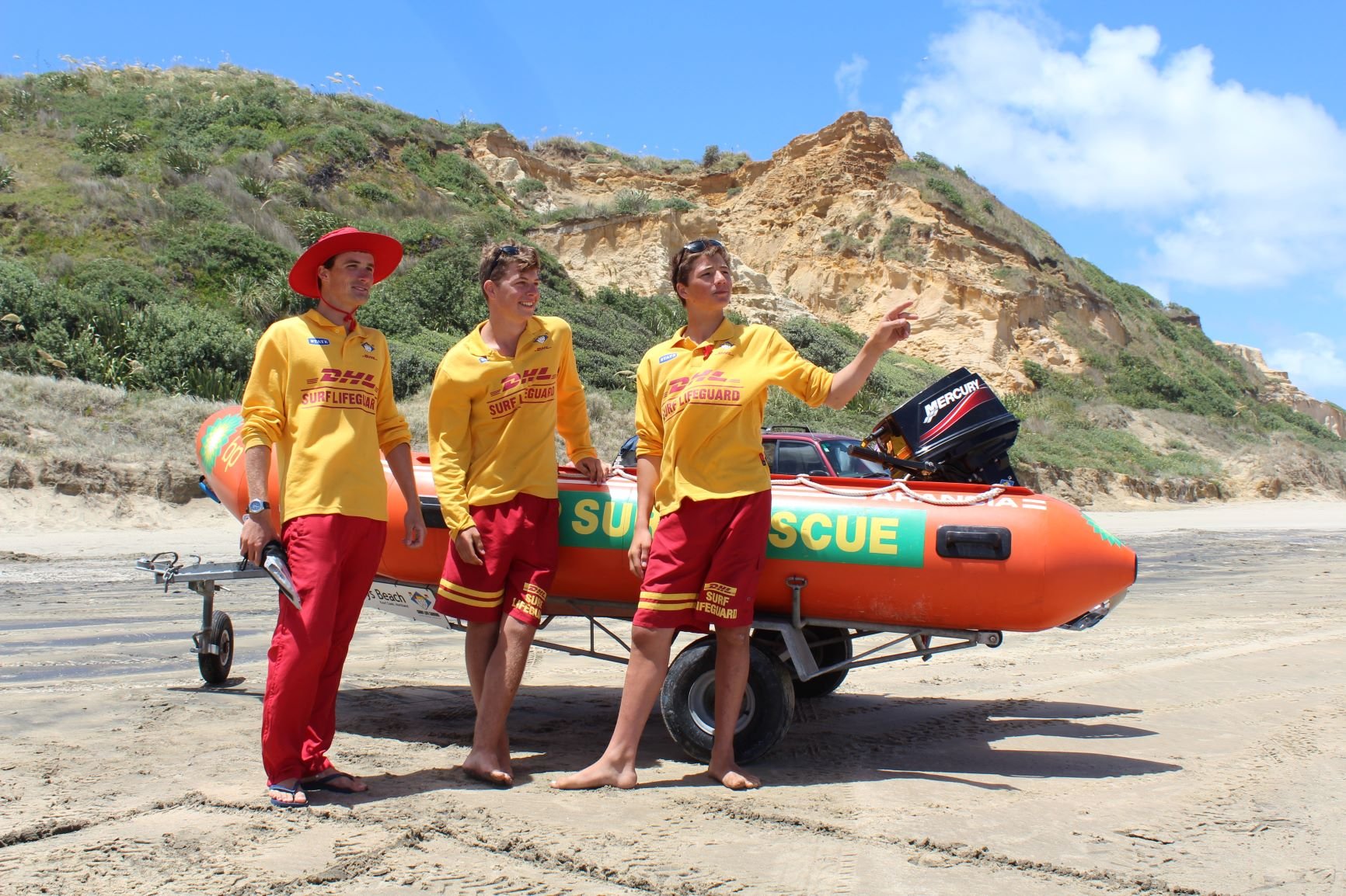 Three surf lifesavers in yellow and red uniforms standing on a sandy beach next to a rescue boat, with green hillside and blue sky in the background