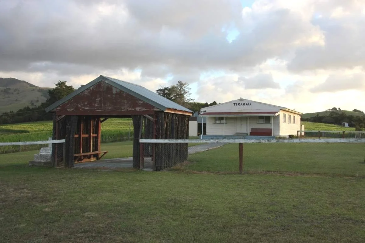 A small white train station building with a sign that reads 'TIRARAU' and a rustic wooden shelter in front, set in a rural landscape with green fields, grassy area, and hills in the background under a partly cloudy sky.
