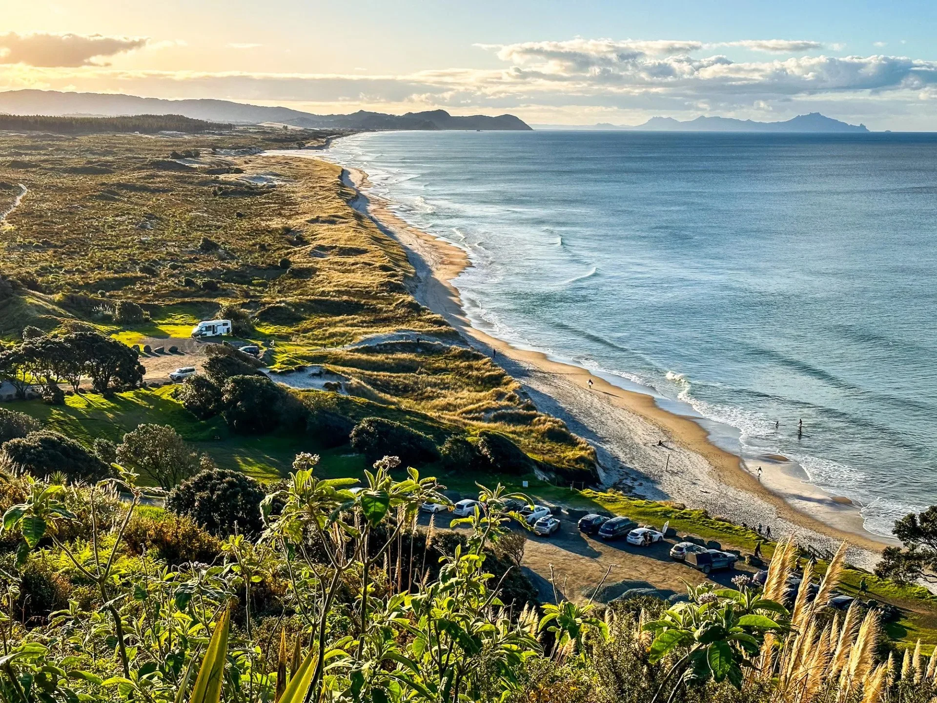 Scenic view of a beach with people surfing, lined with green bushes and parked cars, and a coastal landscape with mountains in the distance under a partly cloudy sky during sunset.