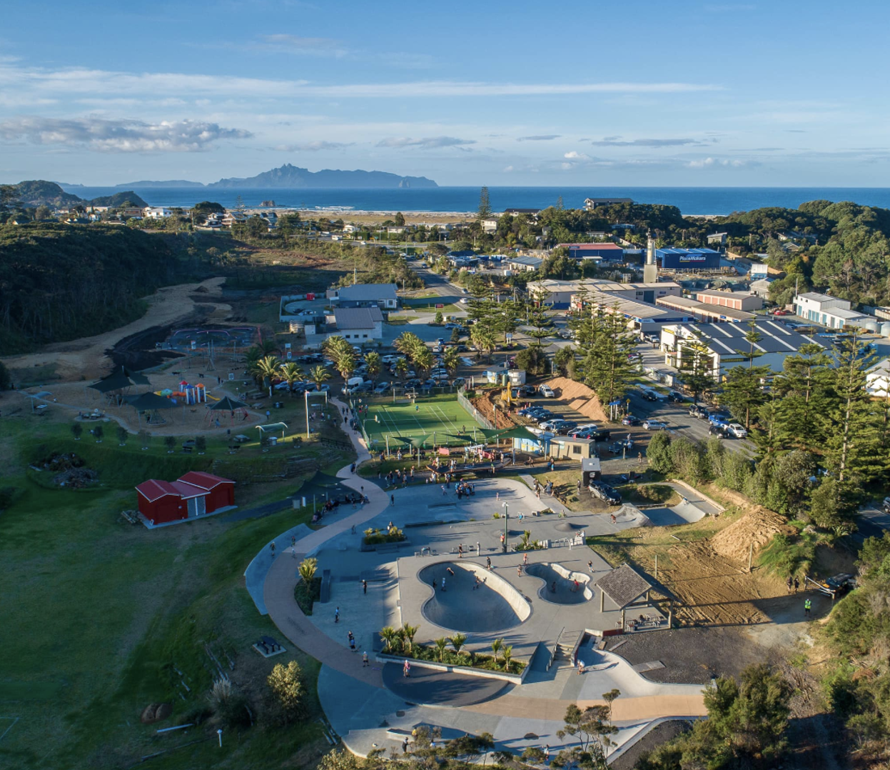 Aerial view of a coastal park with skatepark, playground, and walking paths, surrounded by trees, parking lots, commercial buildings, ocean, and distant mountains.