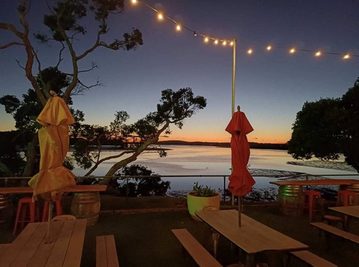 Outdoor patio at sunset with string lights, two closed umbrellas, and wooden tables and benches near a body of water surrounded by trees.