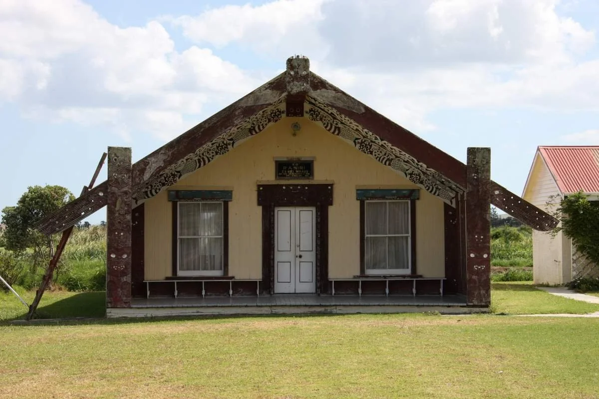 A traditional house with a steep roof decorated with Maori patterns, two front windows with curtains, and a centrally located door, situated on a grassy lawn under a partly cloudy sky.