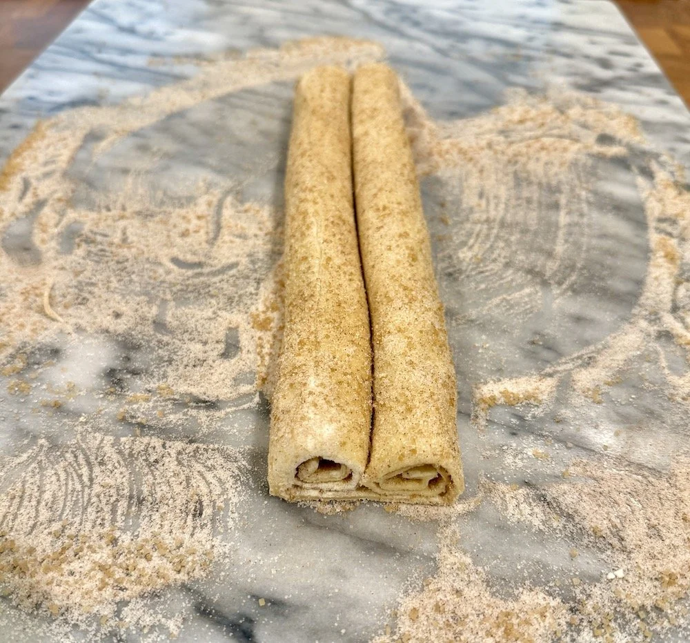 Puff pastry rolled inward from both sides into two tight logs meeting in the center, coated with cinnamon sugar and resting on a marble surface dusted with extra sugar.