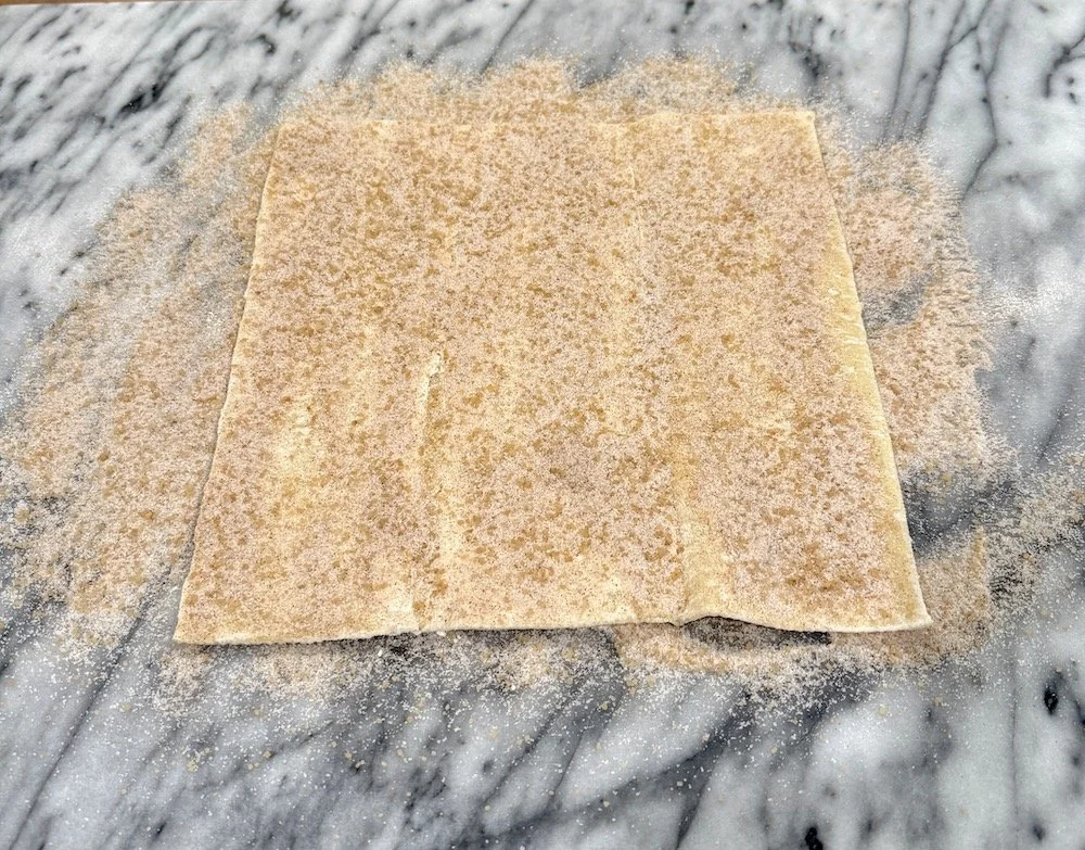 Unfolded sheet of puff pastry on a marble surface, generously sprinkled all over with a cinnamon-sugar mixture.