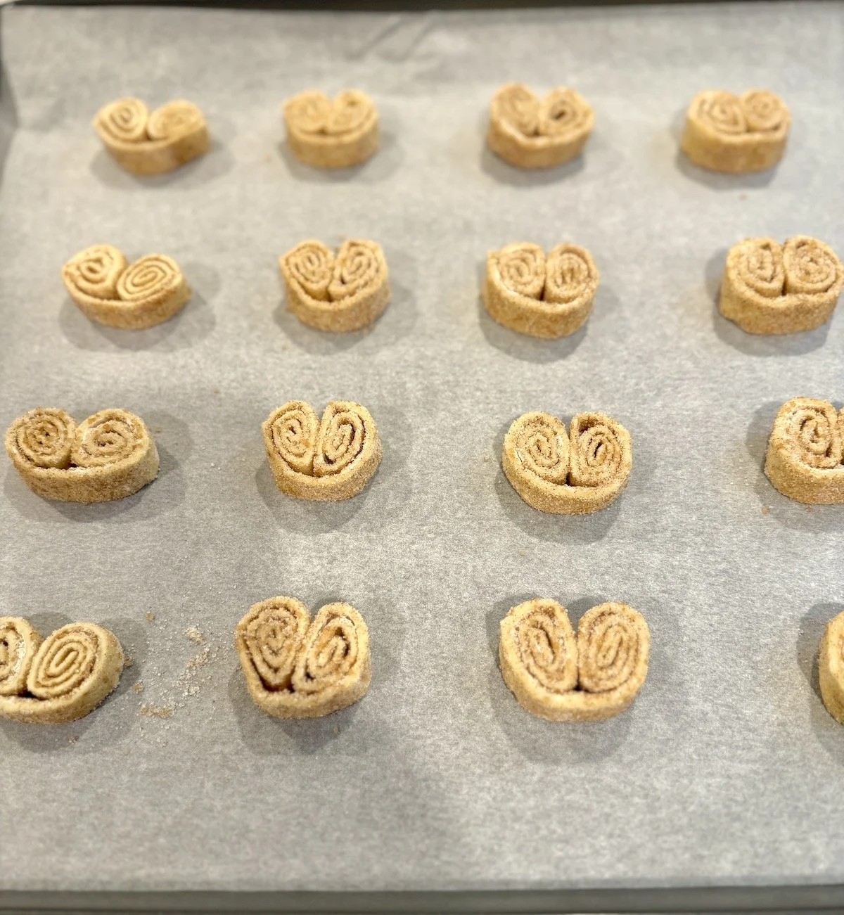 Unbaked cinnamon-sugar palmiers arranged on a parchment-lined baking sheet, showing their signature double-scroll shape and sugared edges.