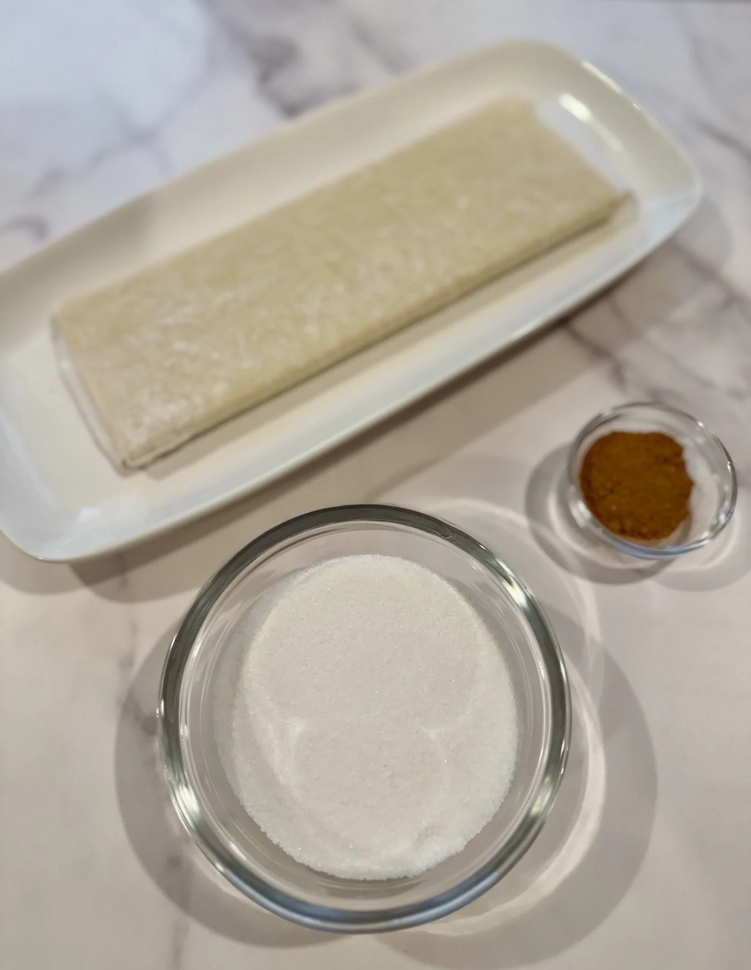 Ingredients for cinnamon sugar palmiers displayed on a marble surface: a folded sheet of puff pastry on a white rectangular plate, a small glass bowl of granulated sugar, and a small glass dish filled with ground cinnamon.