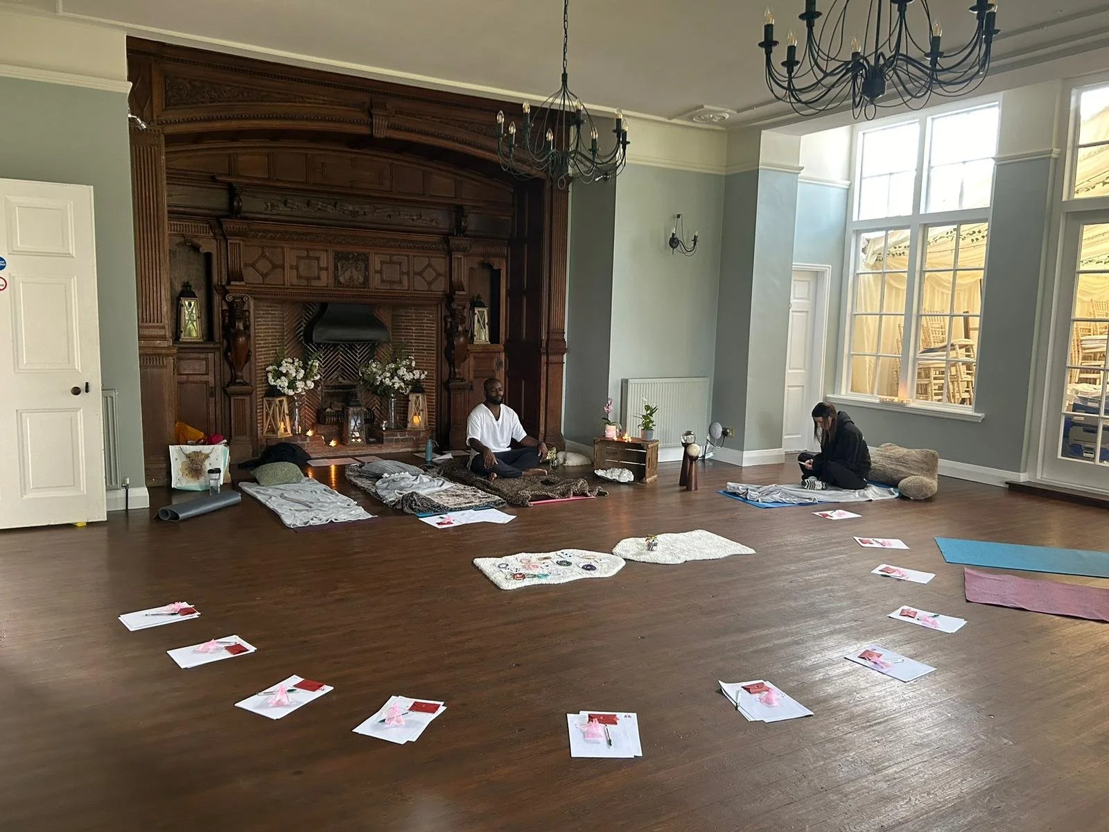 People practicing yoga in a spacious room with large windows, a dark wooden fireplace, and hardwood floors, with yoga mats and instruction sheets laid out on the floor.