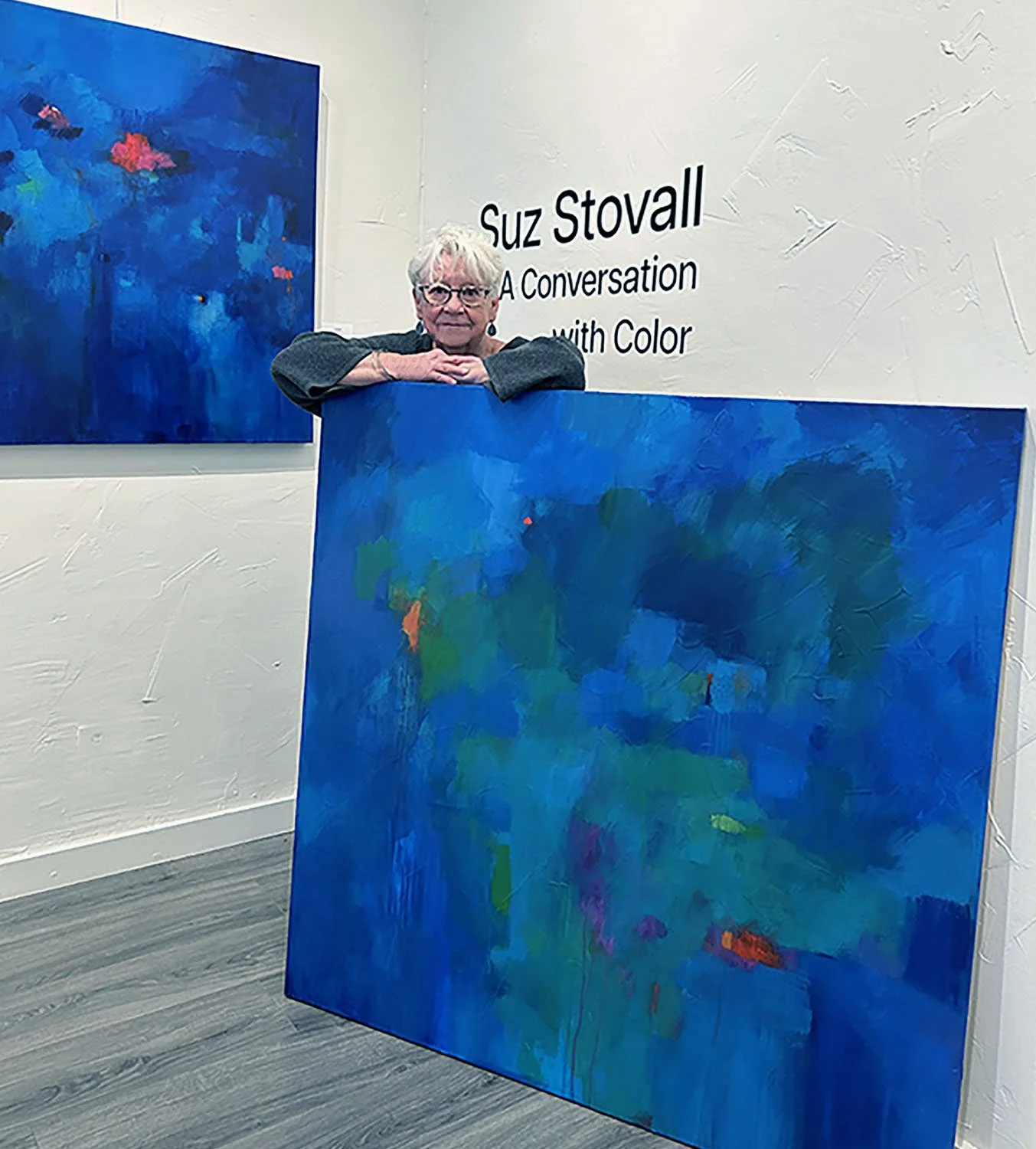 An elderly woman with white hair and glasses leaning on a large, abstract blue painting with green and red accents, inside an art gallery. There are two paintings on the wall, and a sign that reads 'Suz Stovall: A Conversation with Color'.