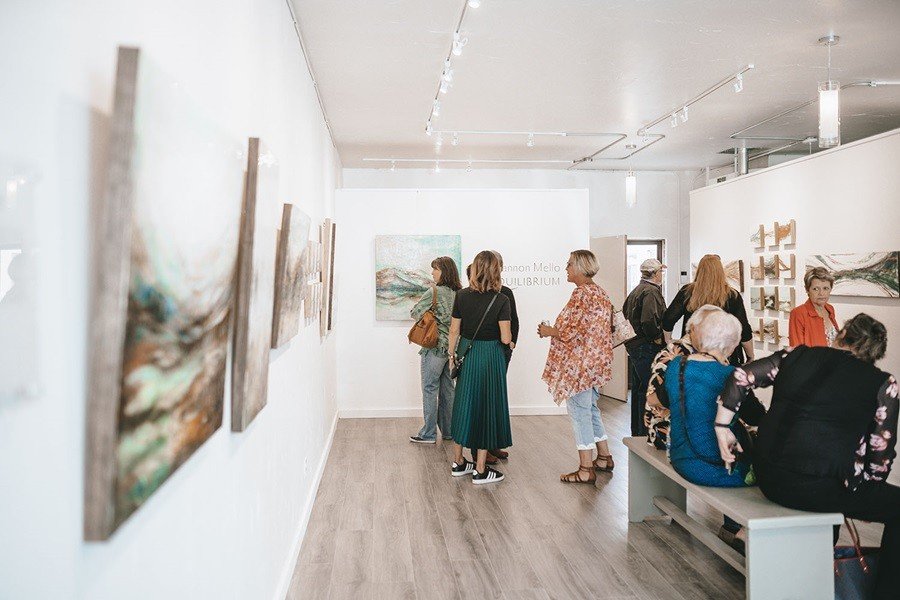 People viewing and discussing artwork in an art gallery with white walls and wooden flooring.