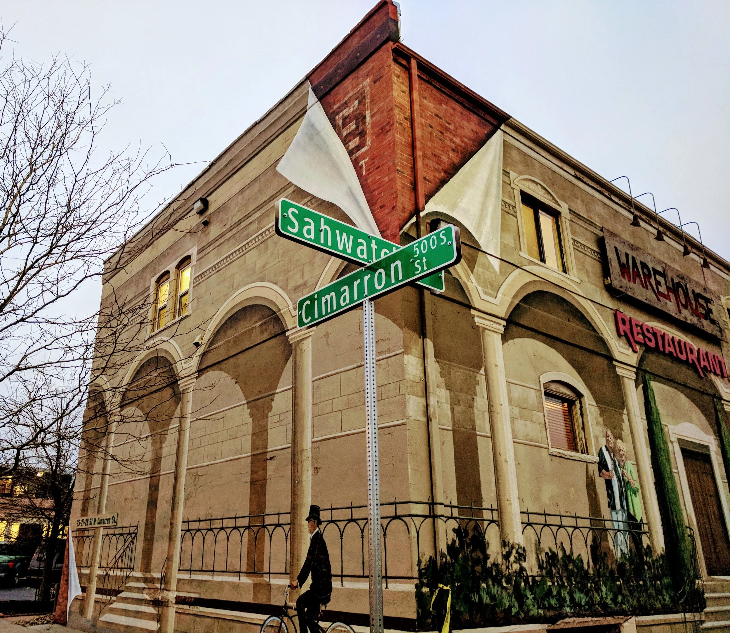 Corner of a building with a brick chimney, decorative stonework, and arched windows. Street signs for Sahwalt and Cameron Streets are visible. There are leafless trees and a person riding a bicycle on the sidewalk.