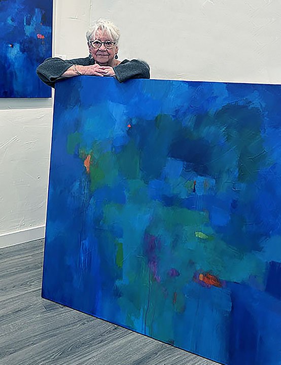 An elderly woman with short white hair and glasses standing behind a large canvas with an abstract blue painting, in an art gallery.