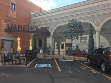 Front view of a building named 'The Wharf House' with a parking lot, including handicapped parking space, and a black vehicle parked on the right side. There are trees and benches near the entrance, and yellow umbrellas are visible in the background.