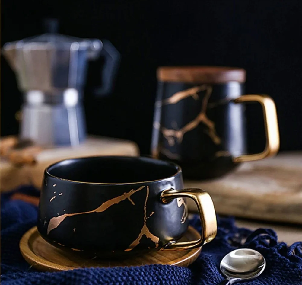 Black coffee cups with gold marble design, a moka pot, and a spoon on a wooden table with dark background.