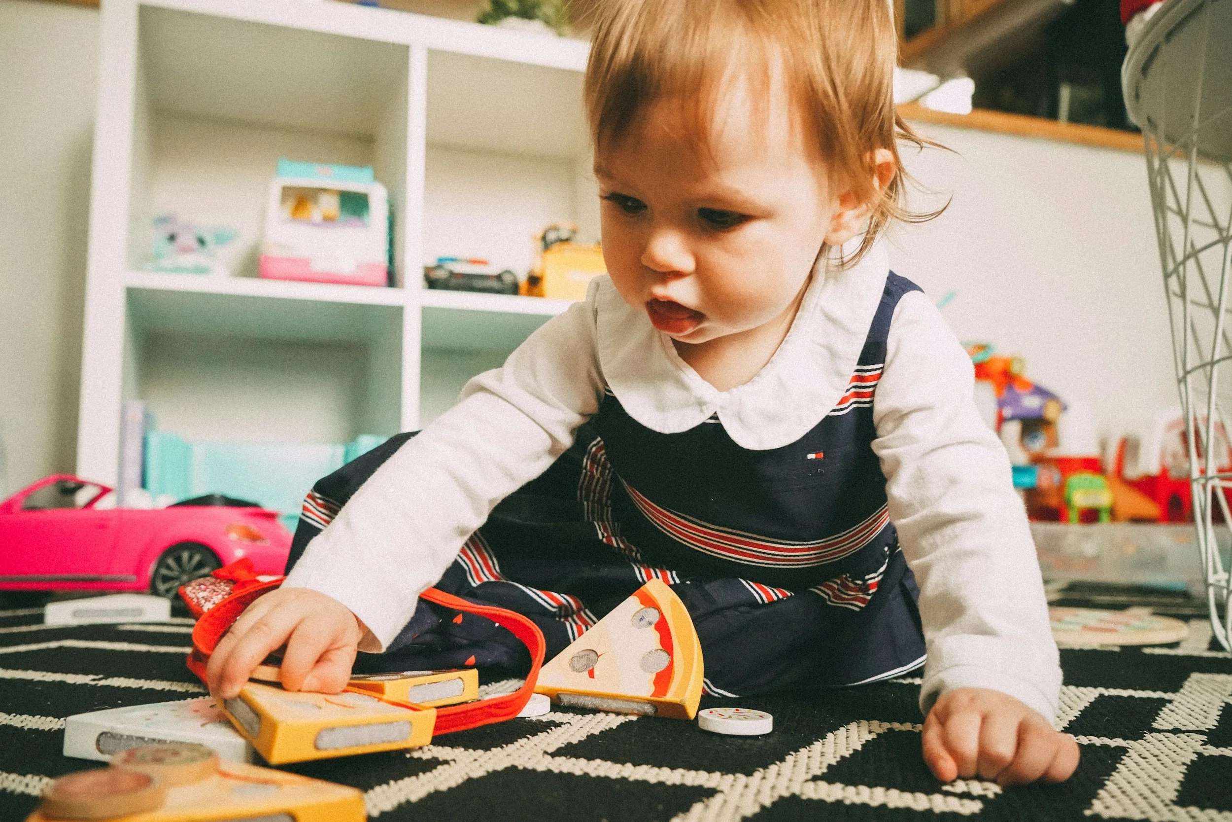 A close-up of an infant sitting on a classroom floor practicing gross motor skills while playing with a pretend plush pizza slice toy.