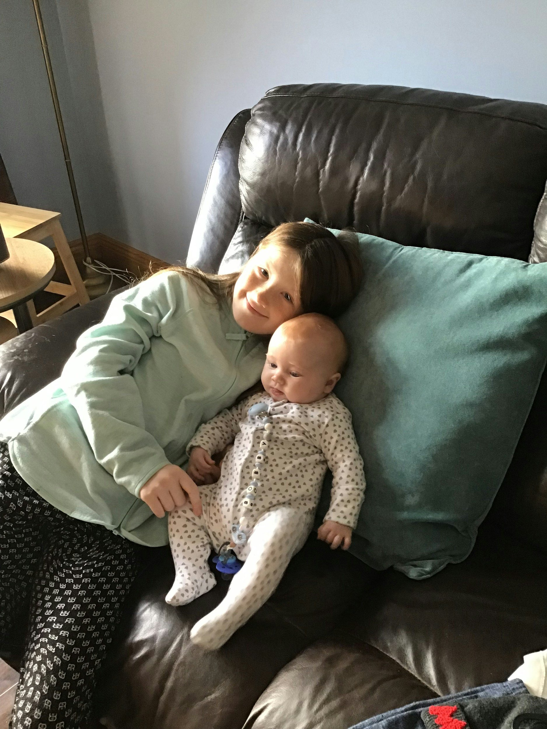 Older child smiling while snuggling a newborn in a chair, practicing early relational connection.