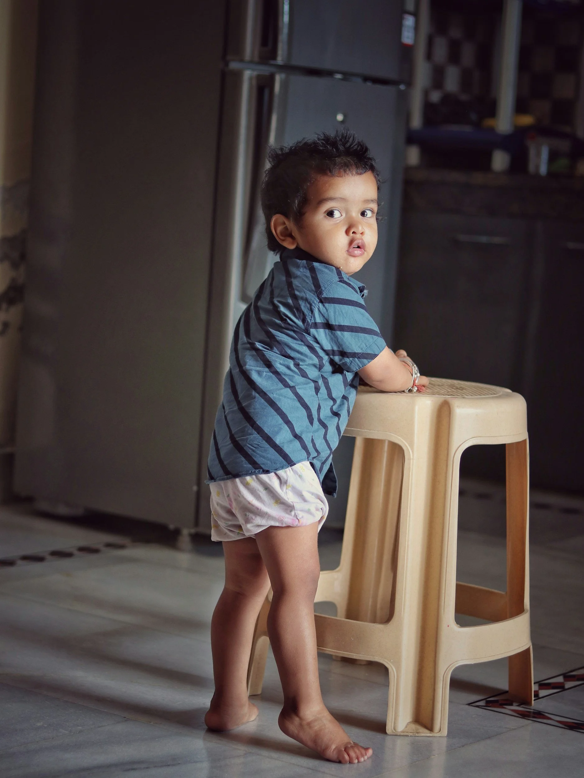 A young child is progressing in his developmental milestones of standing with the support of a chair while looking at the camera.