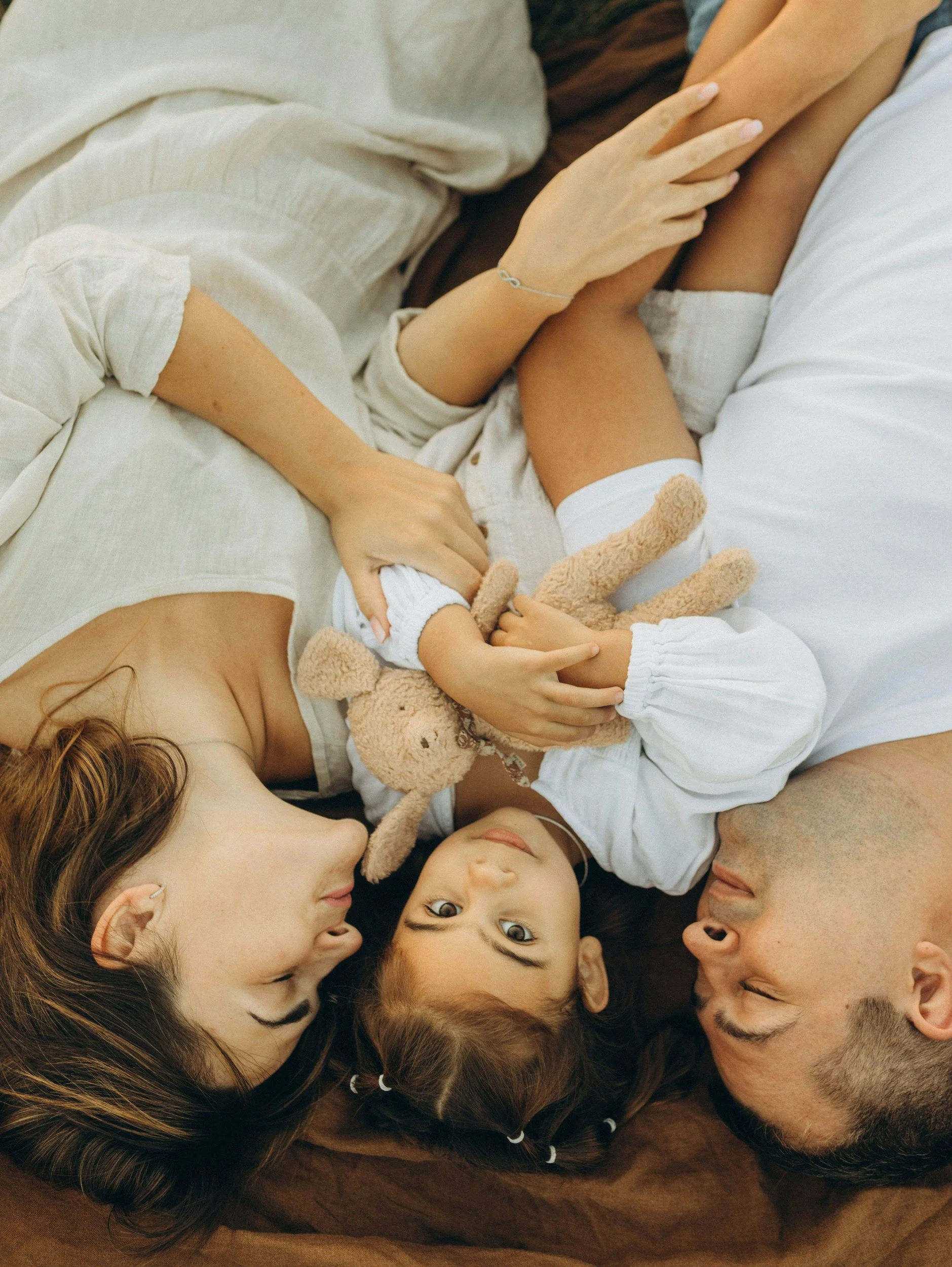 A family lying on a bed, with a mother and a father facing each other and their young child showing joy and connection after postpartum struggles.