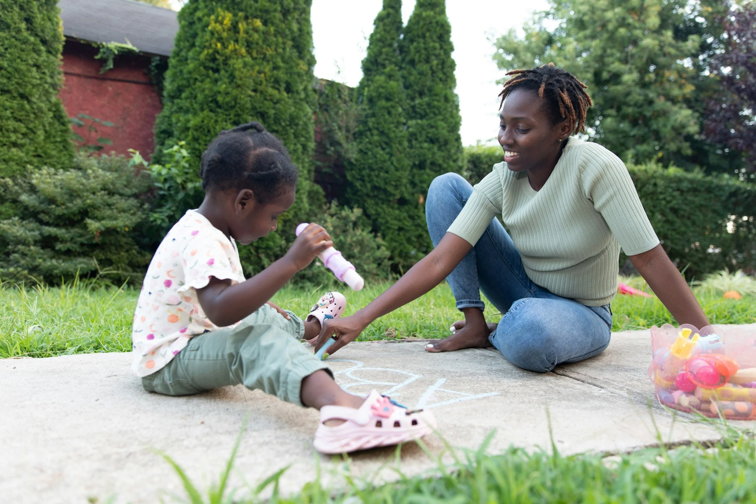 A mother and her preschool-aged child sitting on a driveway outside playing together with colorful sidewalk chalk.
