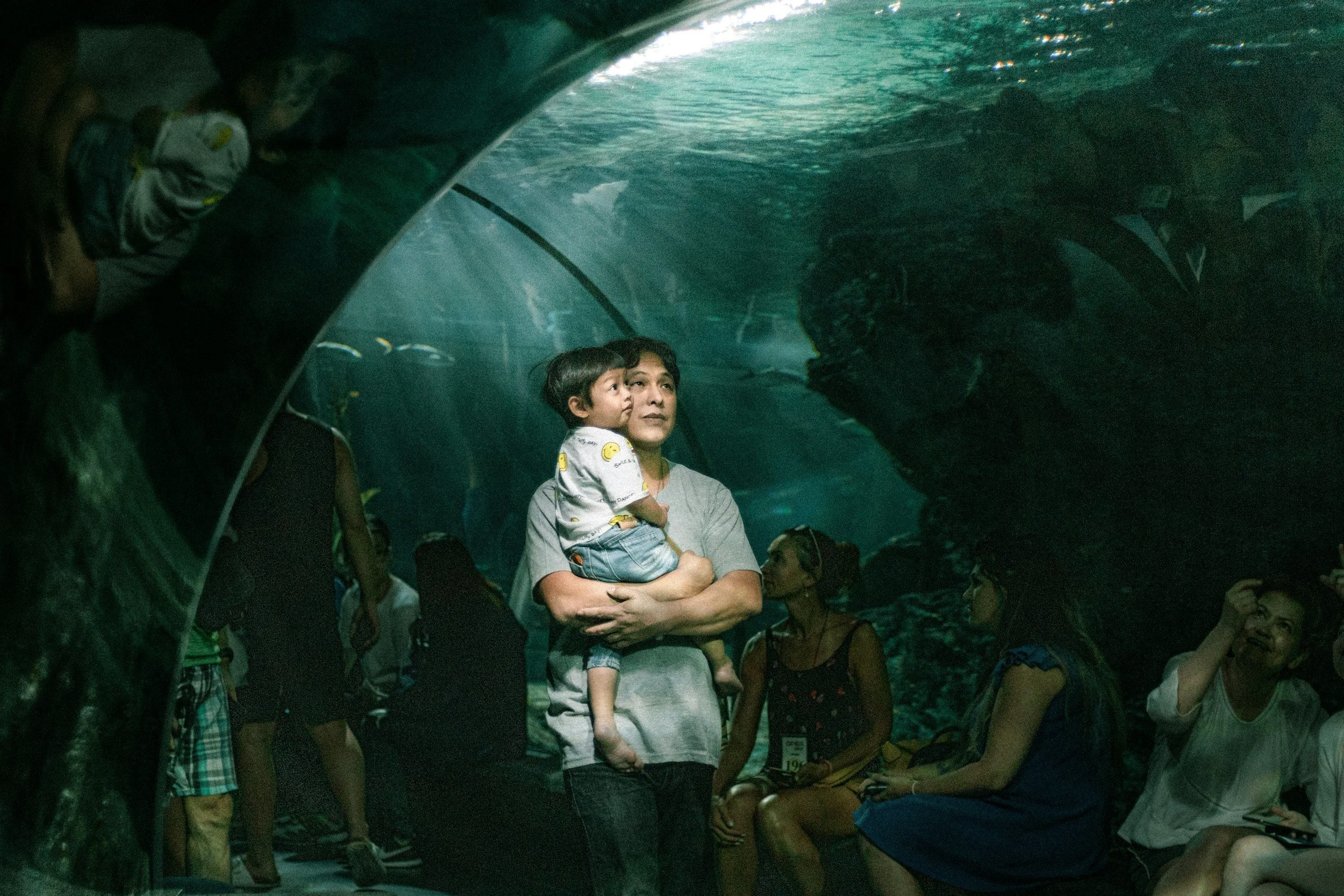 A father holding his toddler while looking at a large blue aquarium tank, modeling curiosity and quiet connection during an early childhood outing.