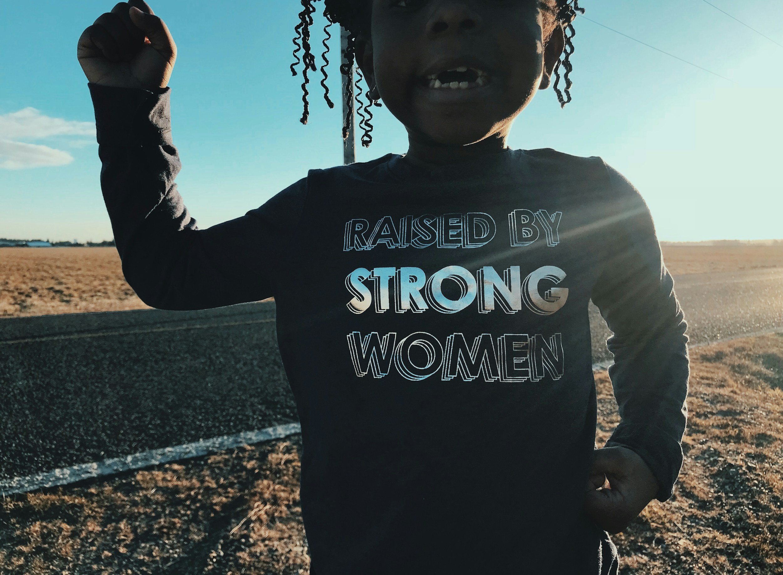 A young girl standing on a sandy beach wearing a shirt that reads 'I am raised by empowered women,' representing the foundation of bravery and identity.